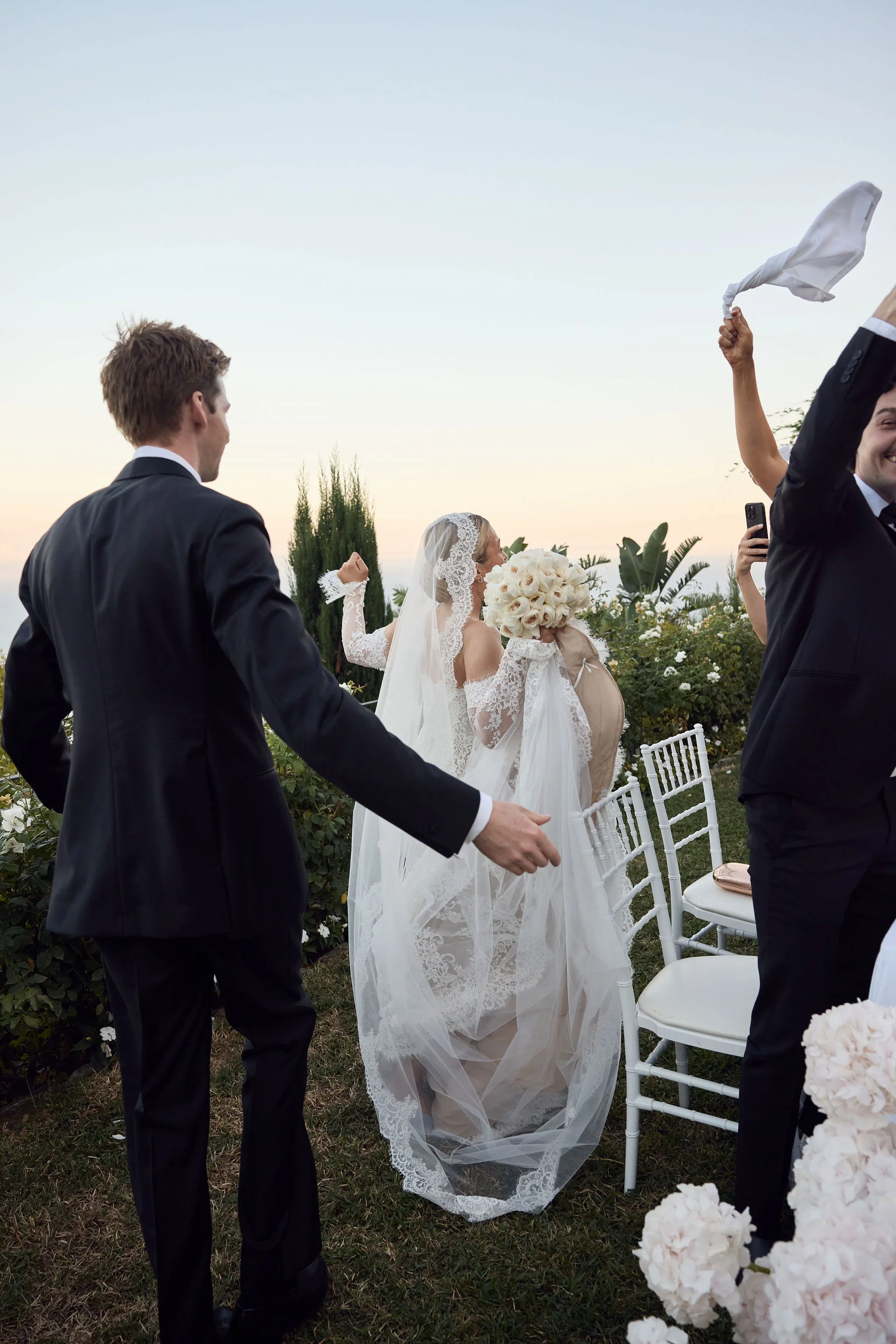 Bride in a lace wedding dress holding a bouquet, surrounded by guests celebrating outdoors during sunset.