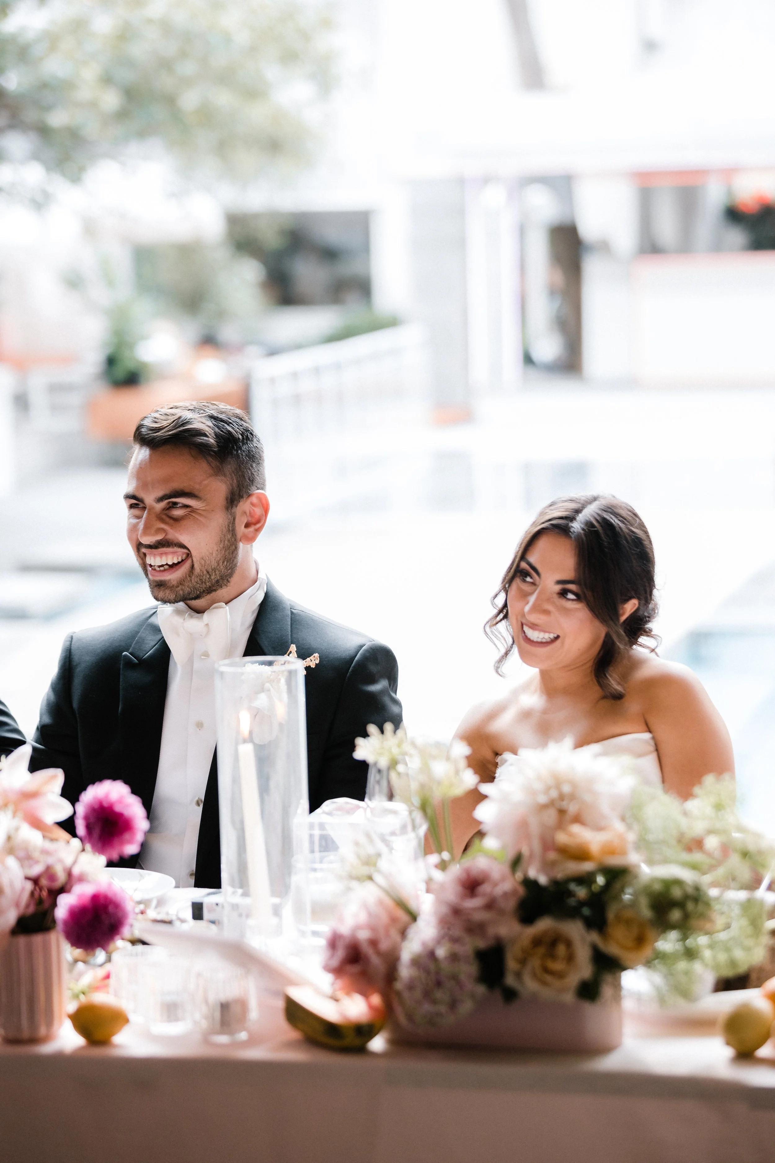 A smiling groom and bride sitting at a wedding reception table with floral centerpieces.