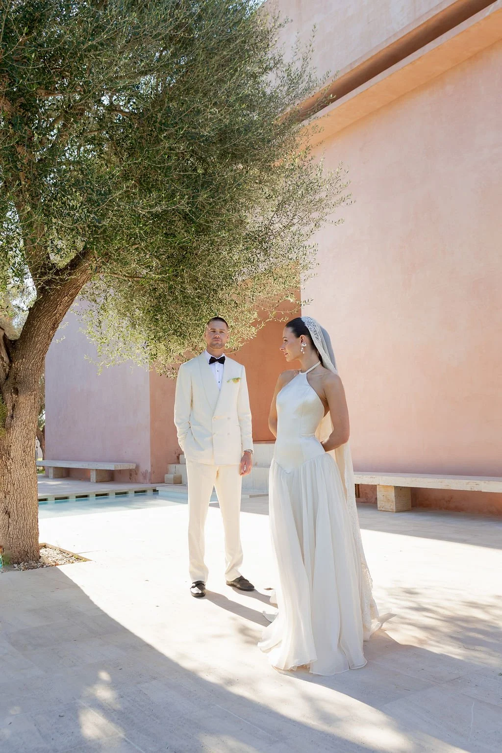 A bride in a white wedding gown and veil and a groom in a white tuxedo with a black bow tie standing outdoors under a tree, with a pinkish wall in the background.