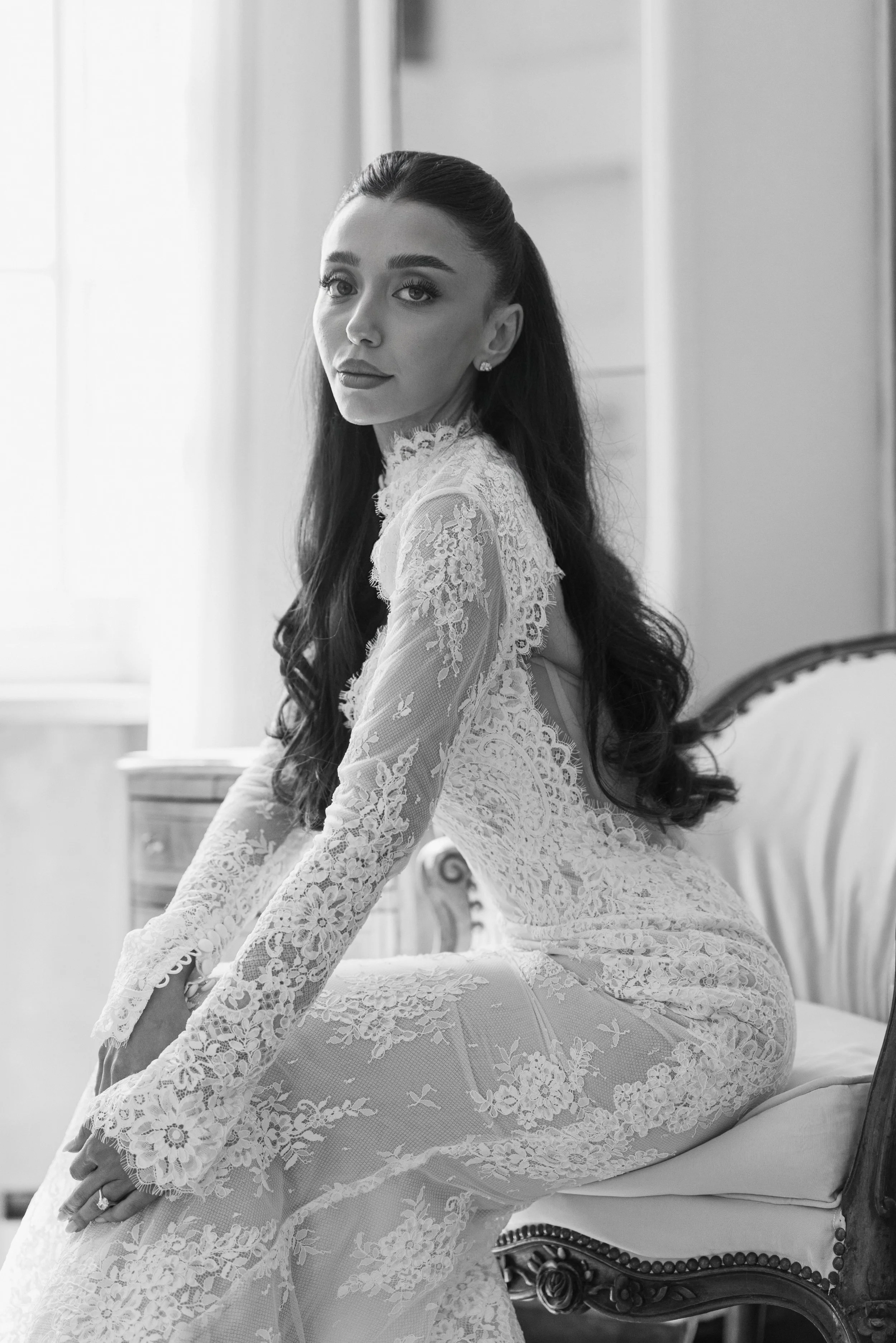 A young woman with long dark hair and earrings, dressed in a lace dress, sitting on a vintage-style chair with detailed woodwork and upholstery, looking at the camera with a composed expression in a softly lit room.