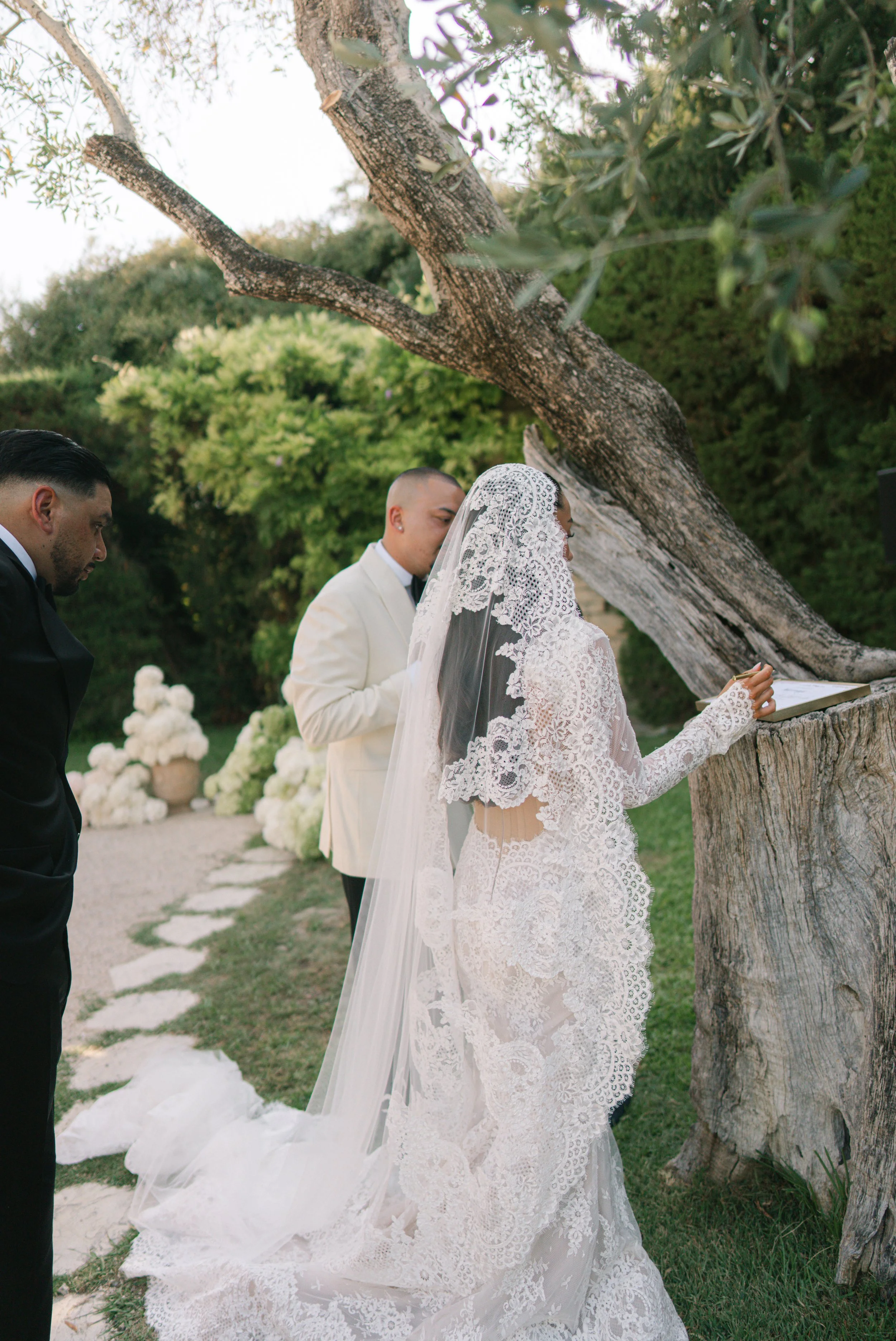 A bride in a lace wedding dress and veil stands outdoors, signing a document next to a groom in a white tuxedo, with a man in a black tuxedo beside her, under a large tree.