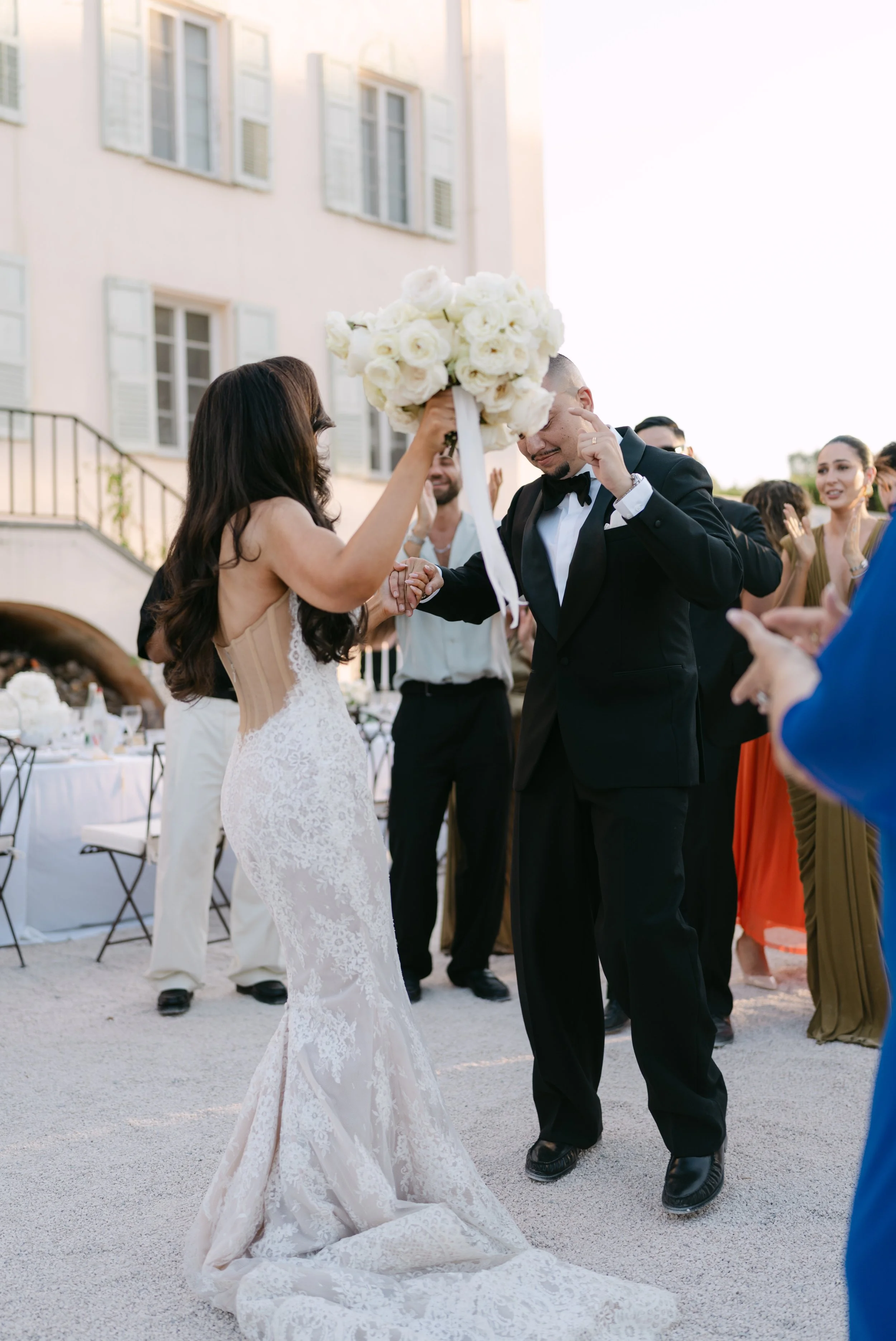 A bride and groom hold hands during their wedding ceremony outdoors, with guests clapping and celebrating in the background near a light-colored building with open windows.