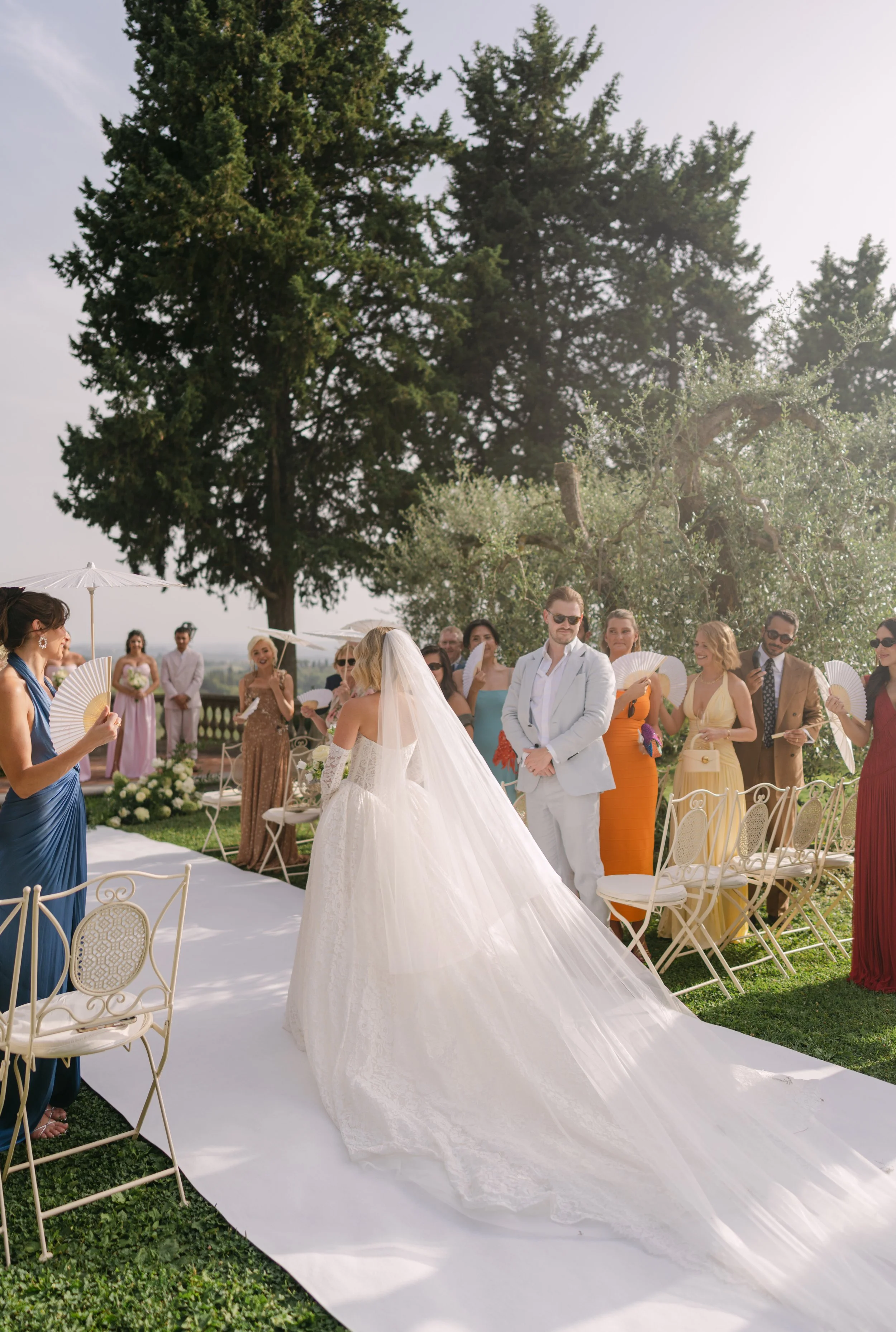 A bride in a white wedding gown and veil walking down an outdoor aisle at a wedding ceremony, surrounded by seated guests and standing people holding fans, with large trees and a bright sky in the background.