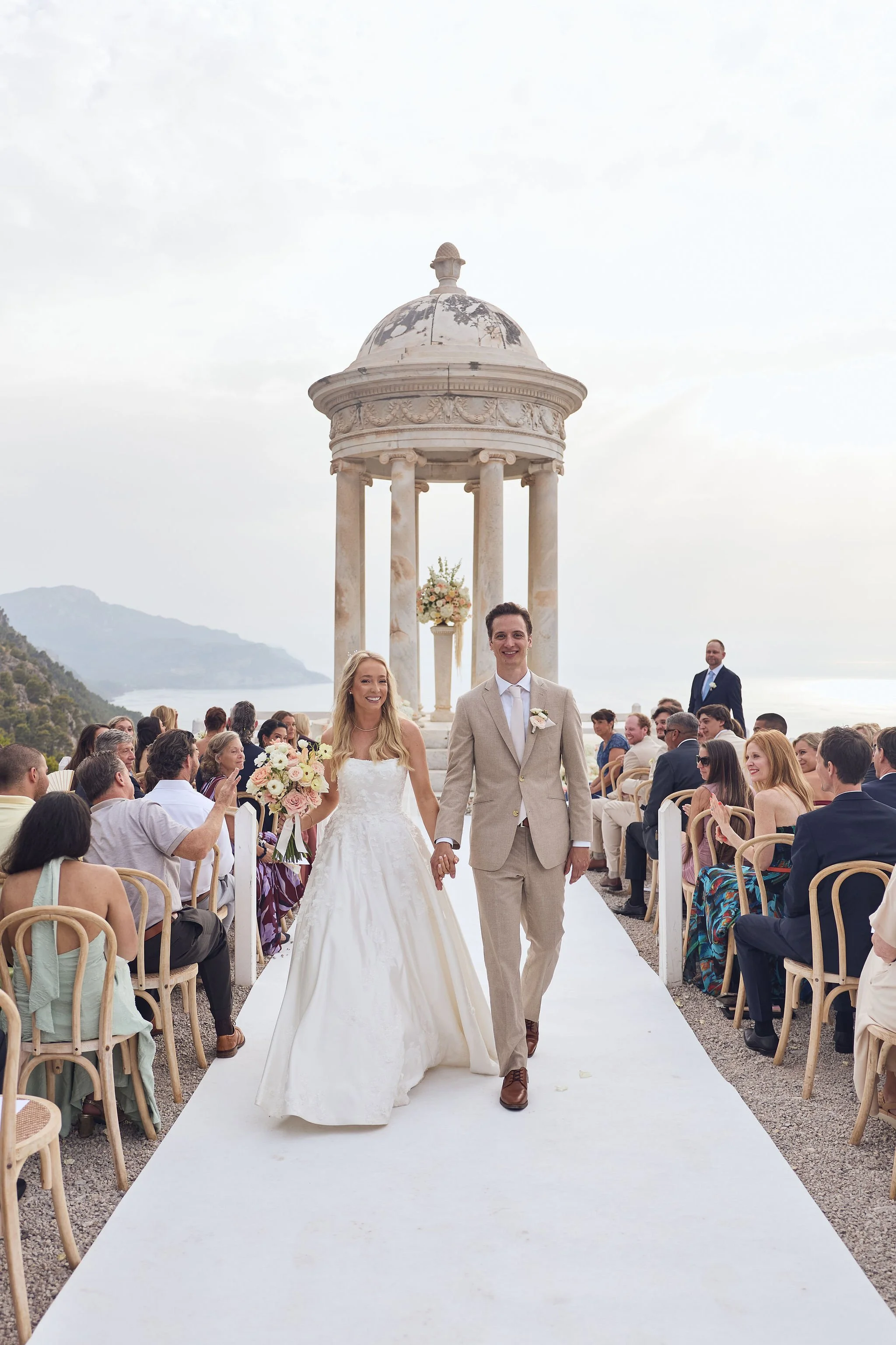 A bride and groom walking down the aisle at an outdoor wedding ceremony with guests seated on either side, a white aisle runner, and a stone pavilion in the background overlooking a coastline.