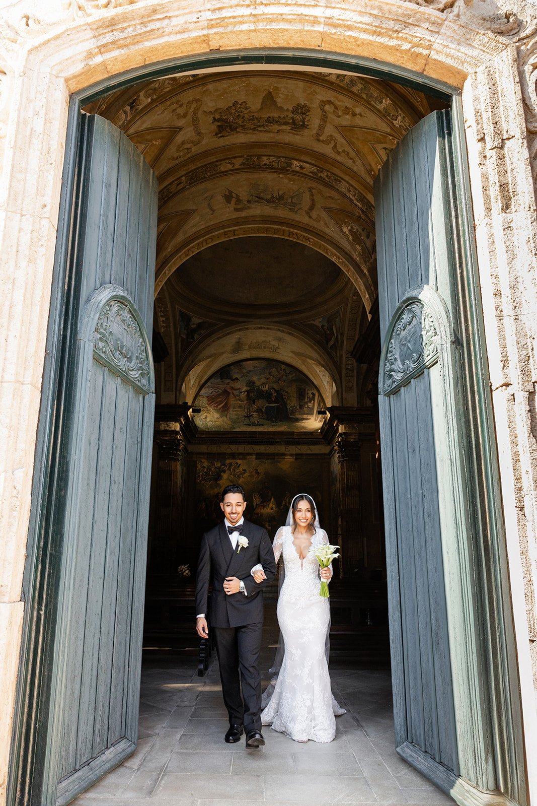 A bride and groom walking out of a church through large open green doors, smiling. The groom wears a black tuxedo with a bow tie and white boutonniere. The bride wears a white lace wedding dress and holds a bouquet of white lilies. The church has hig