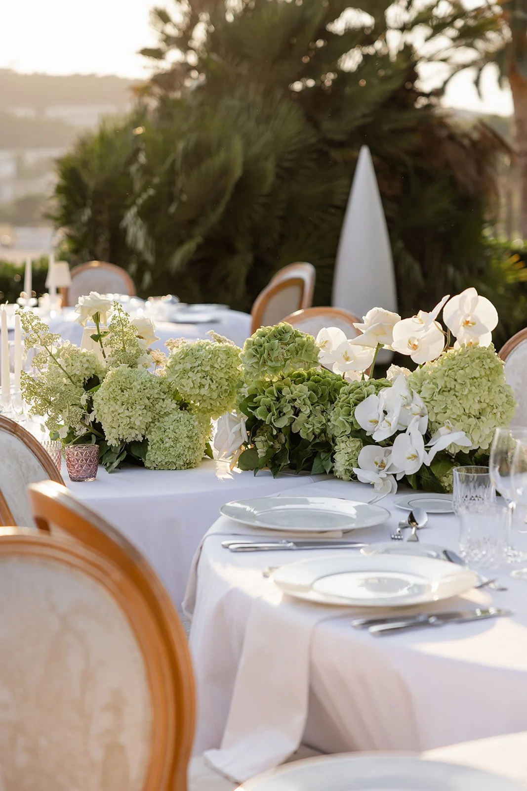 Elegant outdoor dining table decorated with white and green floral arrangements, set with white plates, silverware, and glassware on a white tablecloth, with chairs around it and lush greenery in the background.