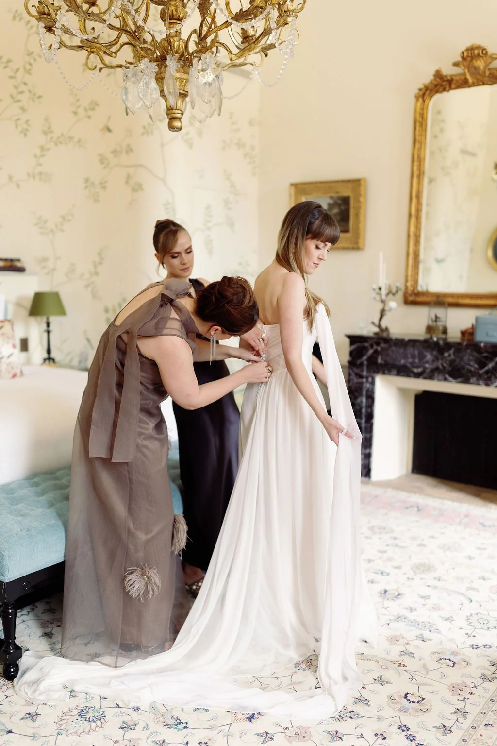 A bride in a white wedding gown standing in a decorated room with ornate gold mirrors and chandelier, while two women assist her with her dress.