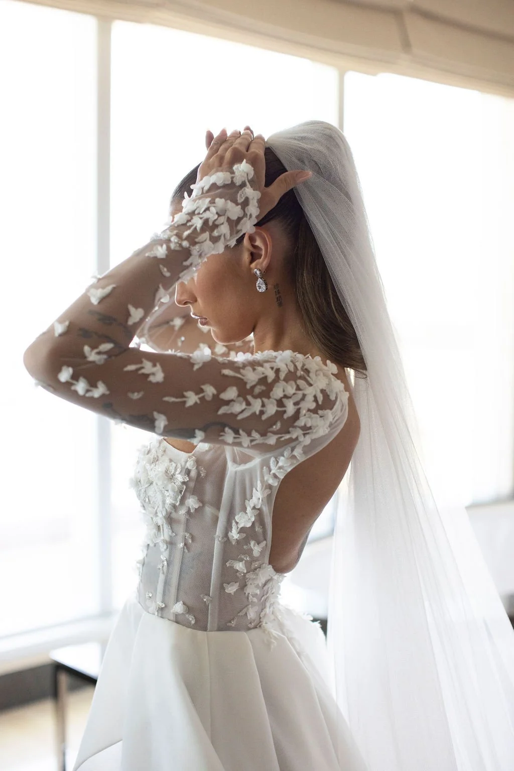 Bride wearing a lace wedding dress and veil adjusting her hair beside large windows.