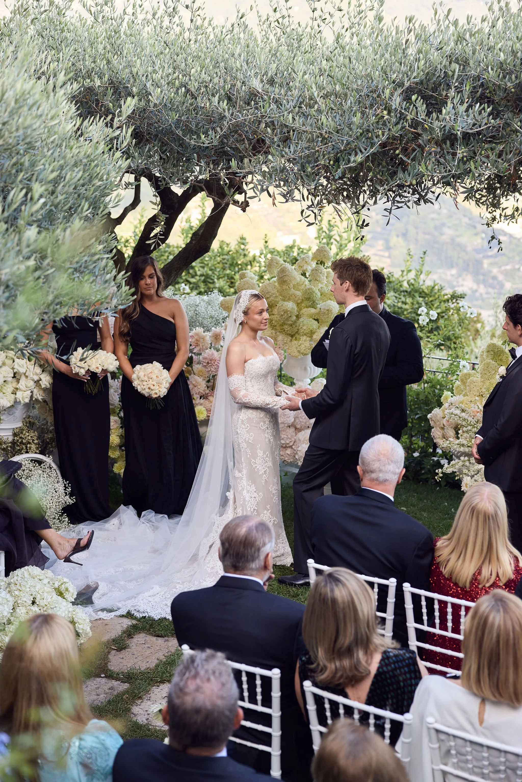 A wedding ceremony is taking place outdoors under a large tree, with the bride and groom holding hands and exchanging vows. The bride is wearing a white lace wedding dress with long lace sleeves and a veil, while the groom is in a black tuxedo. Bride