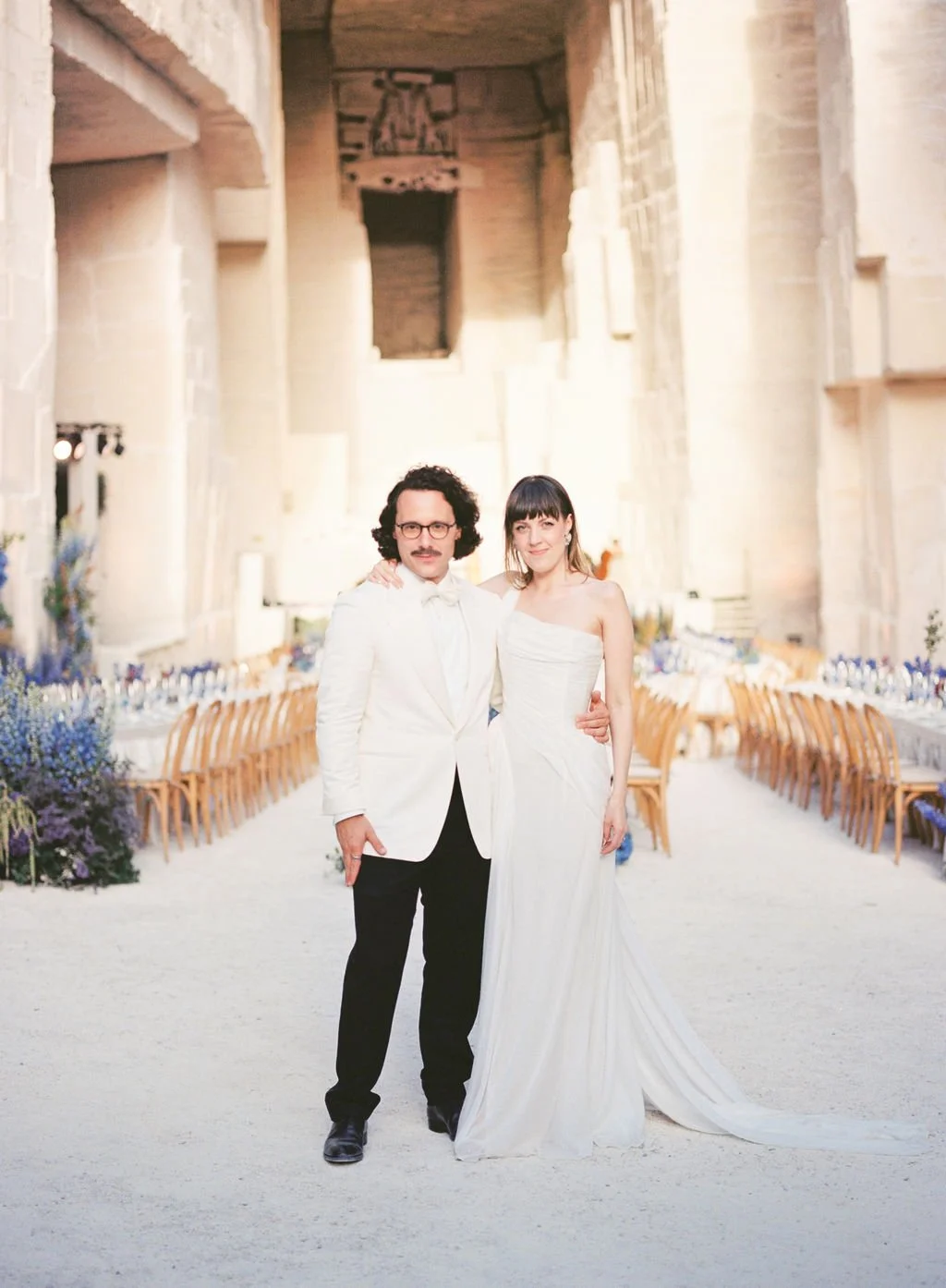 A man and woman in wedding attire standing inside a grand, historic hall with high ceilings and stone walls, decorated for a wedding reception.