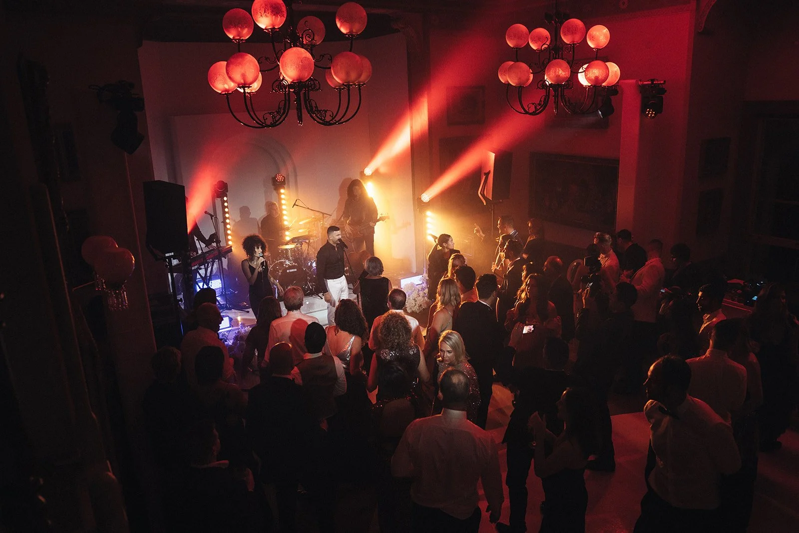 People dancing and enjoying a live music performance in a dimly lit venue with colorful stage lighting and chandeliers.