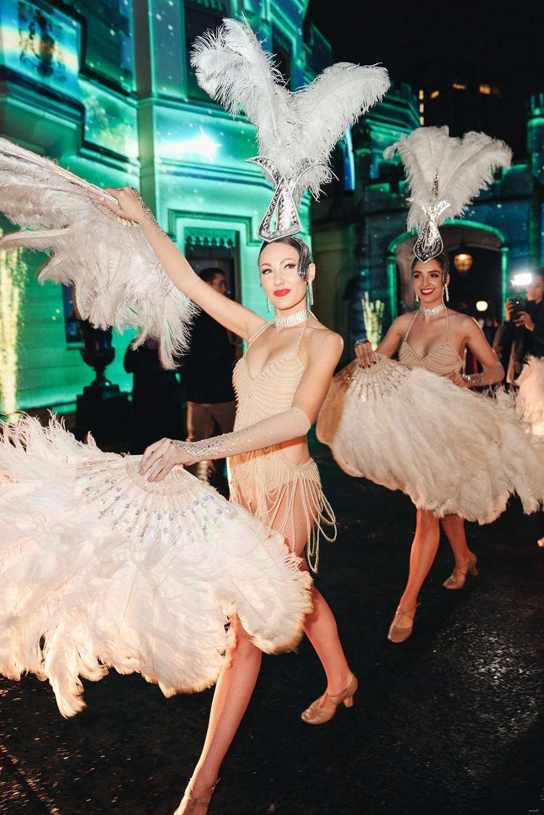 Two women in sparkling costumes with feathered headpieces performing a dance at a nighttime event, with illuminated buildings in the background.