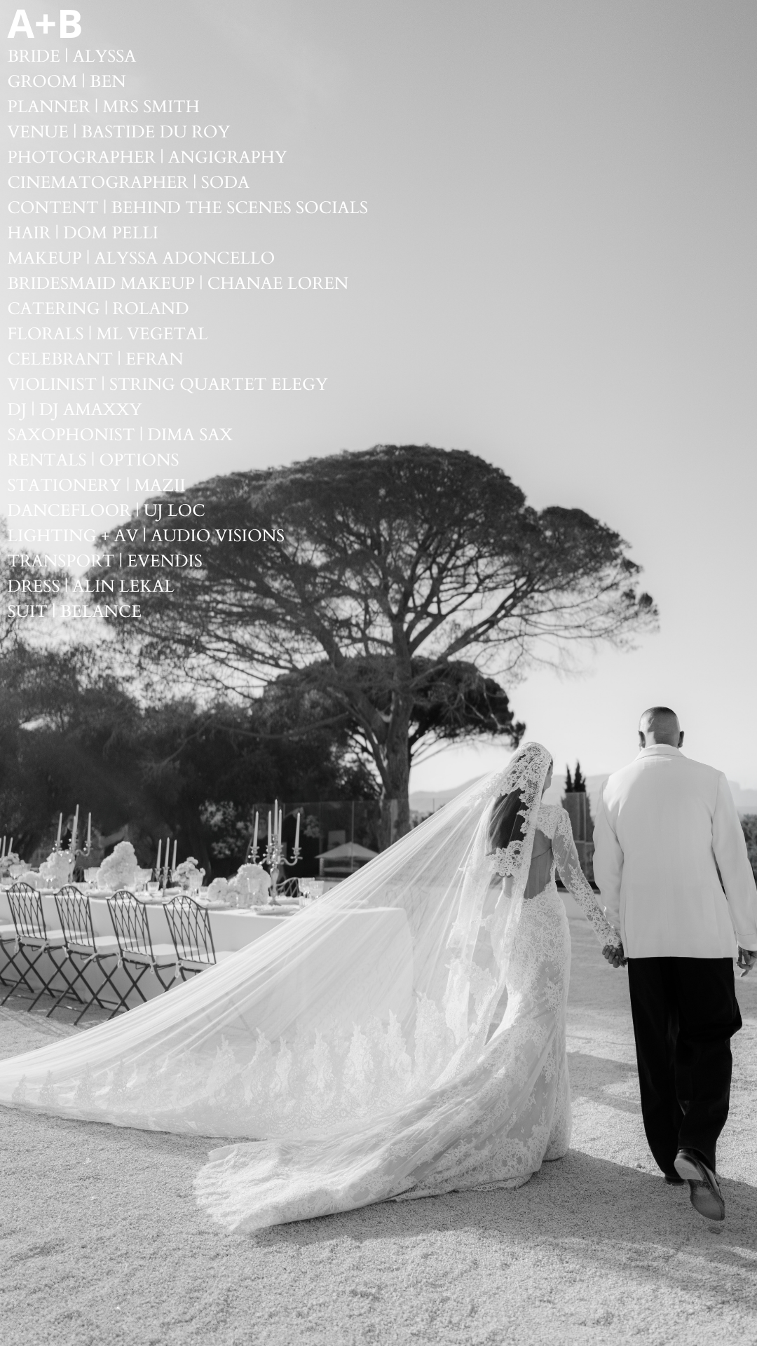 A couple, a bride in a wedding gown with a long train and veil and a groom in a white suit, walk hand in hand outdoors under a large tree. A decorated table with candles and floral arrangements is set up nearby, suggesting a wedding celebration.