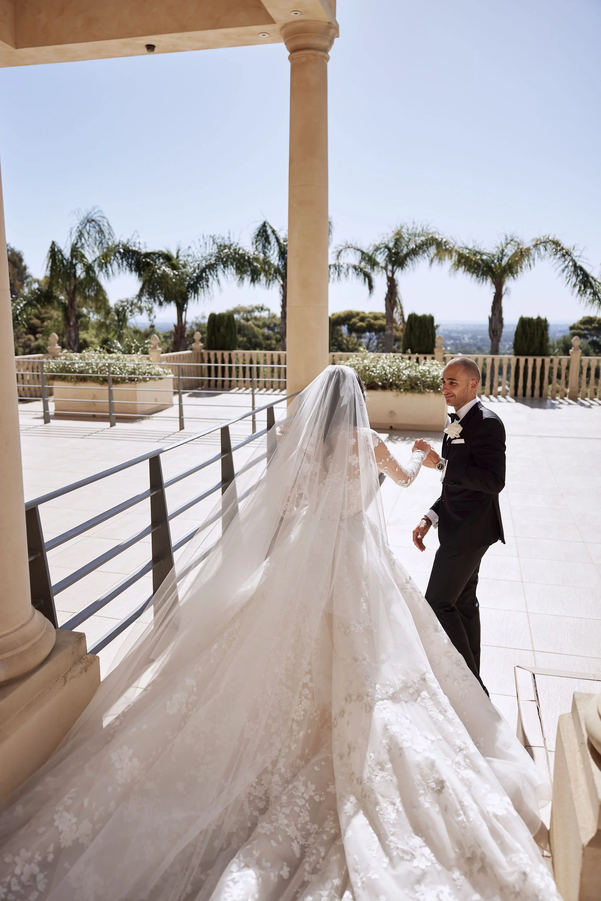 Bride and groom holding hands on a terrace with palm trees in the background.