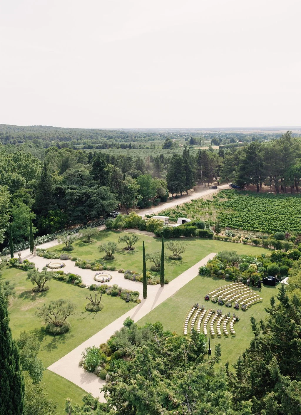 A scenic view of a garden on a sunny day, featuring a well-maintained lawn, small trees, winding pathways, and rows of arranged chairs, with a backdrop of forested hills and open fields.