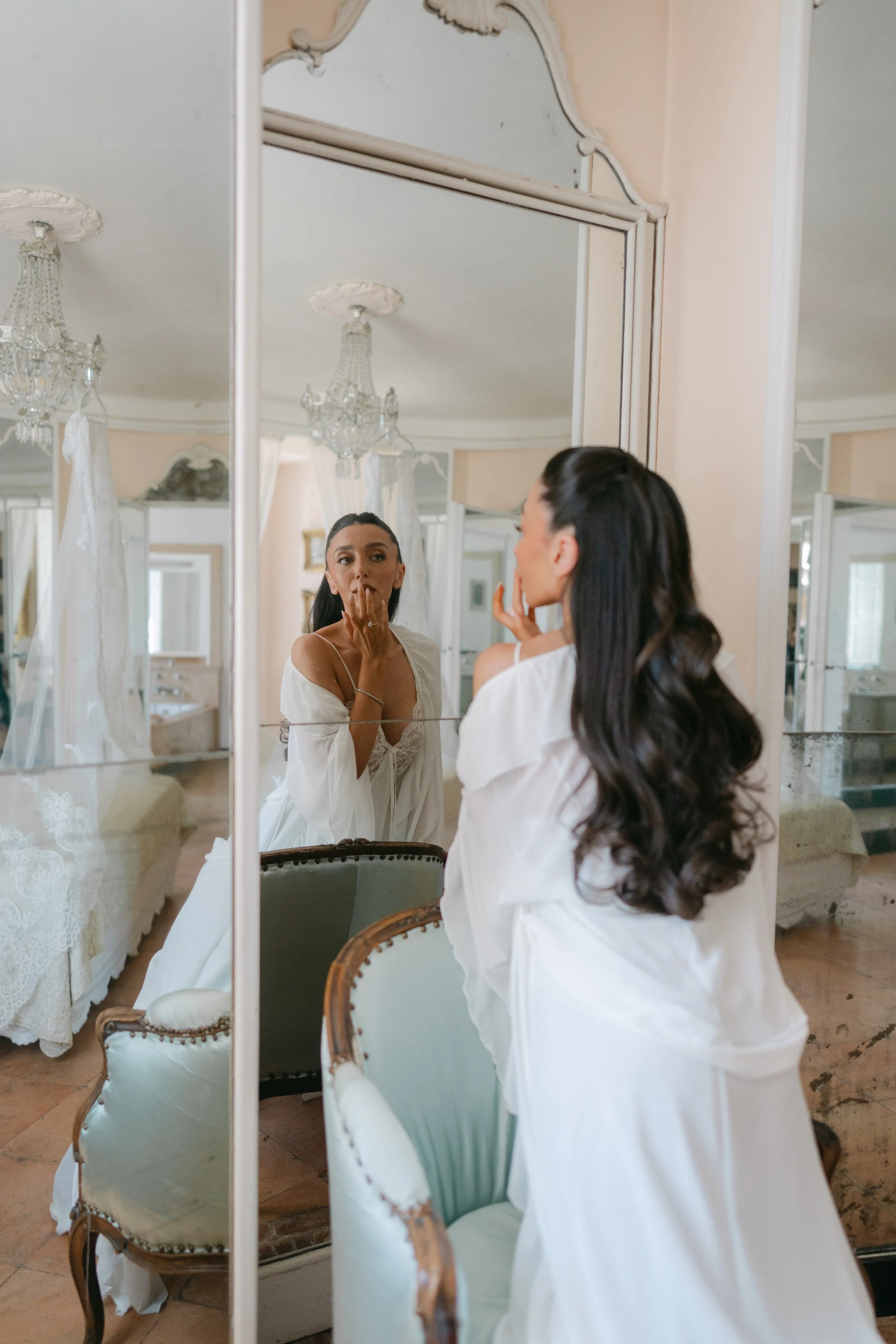 A woman in a white robe looking into a mirror and touching her lips, with a reflection showing her face, in a bedroom decorated with chandeliers and vintage furniture.