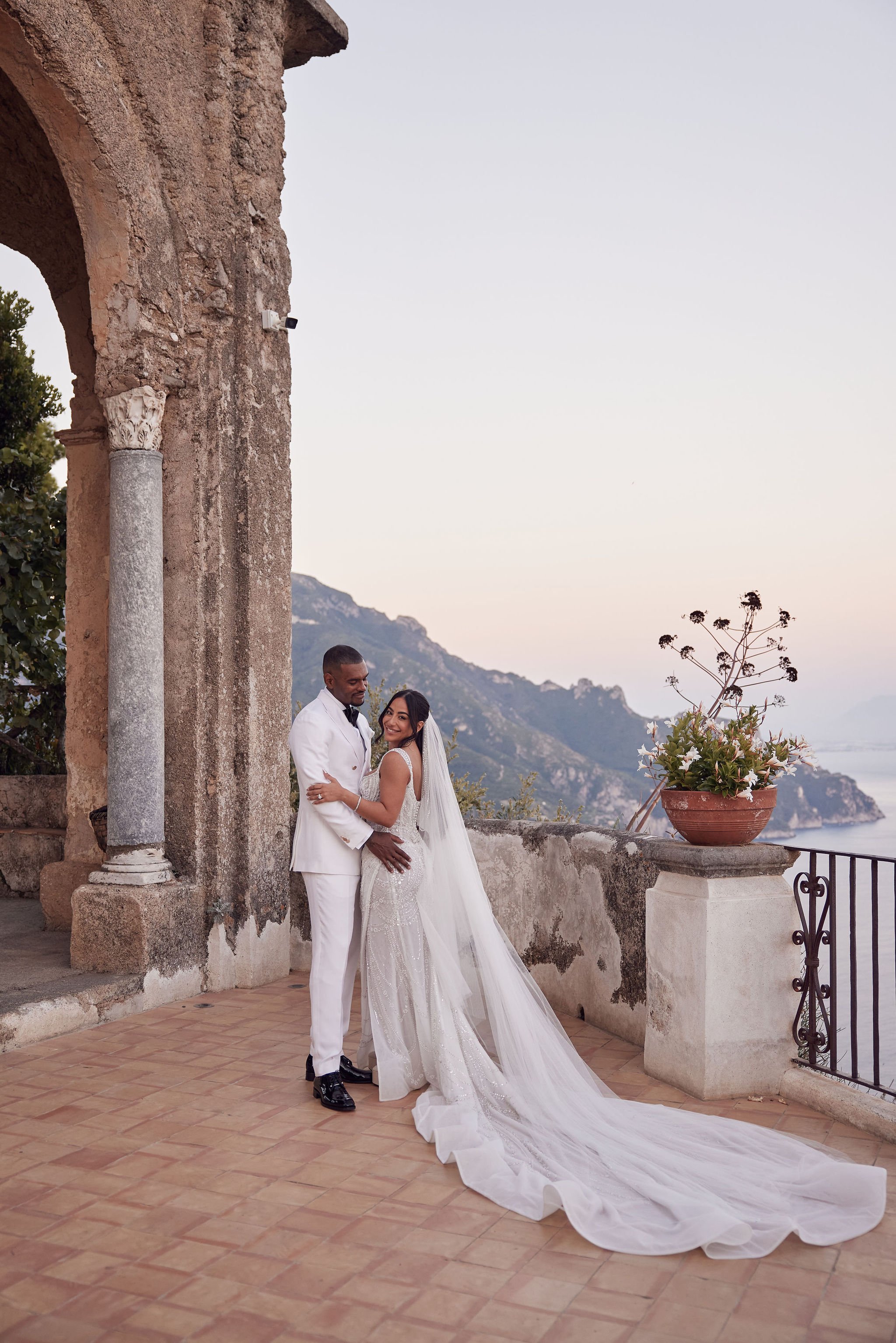 A bride and groom standing together outdoors on a terrace with a scenic mountain and sea view in the background. The bride is wearing a white gown with a long train and veil, and the groom is dressed in a white suit. They are smiling and embracing each other.