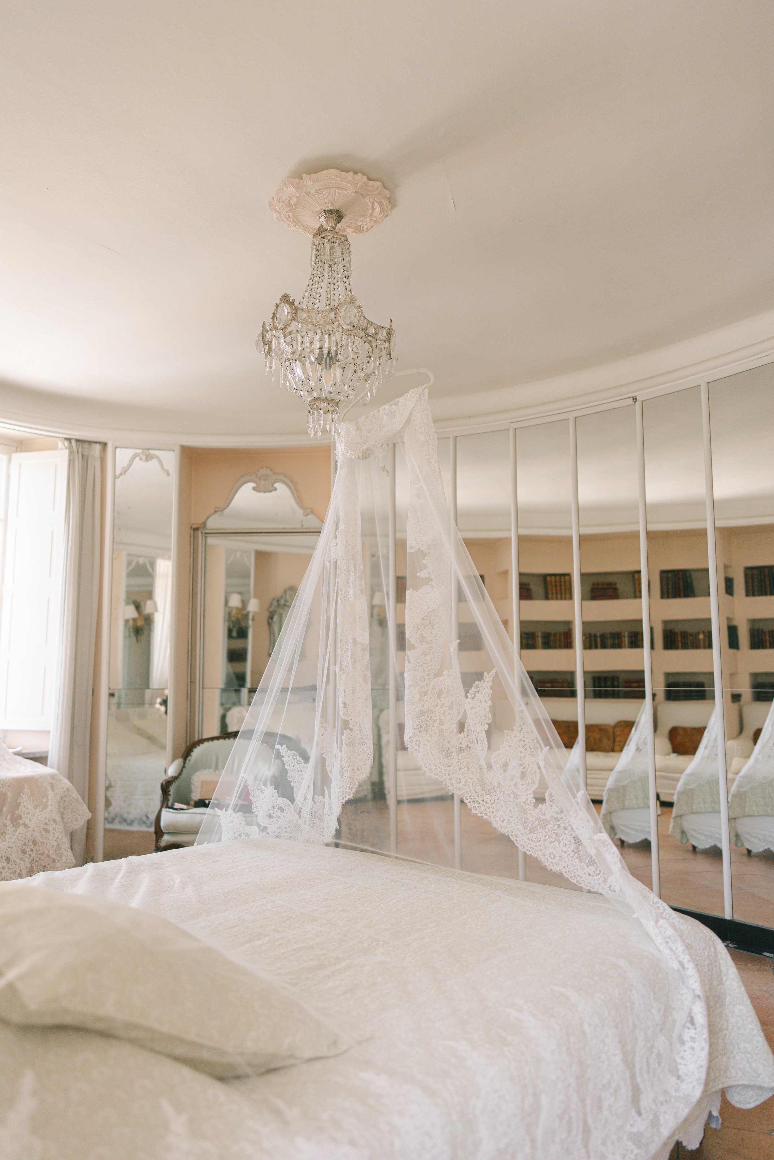 A luxurious room with a chandelier hanging from a decorated ceiling, a canopy bed with lace curtains, a large mirror, and bookshelves in the background.