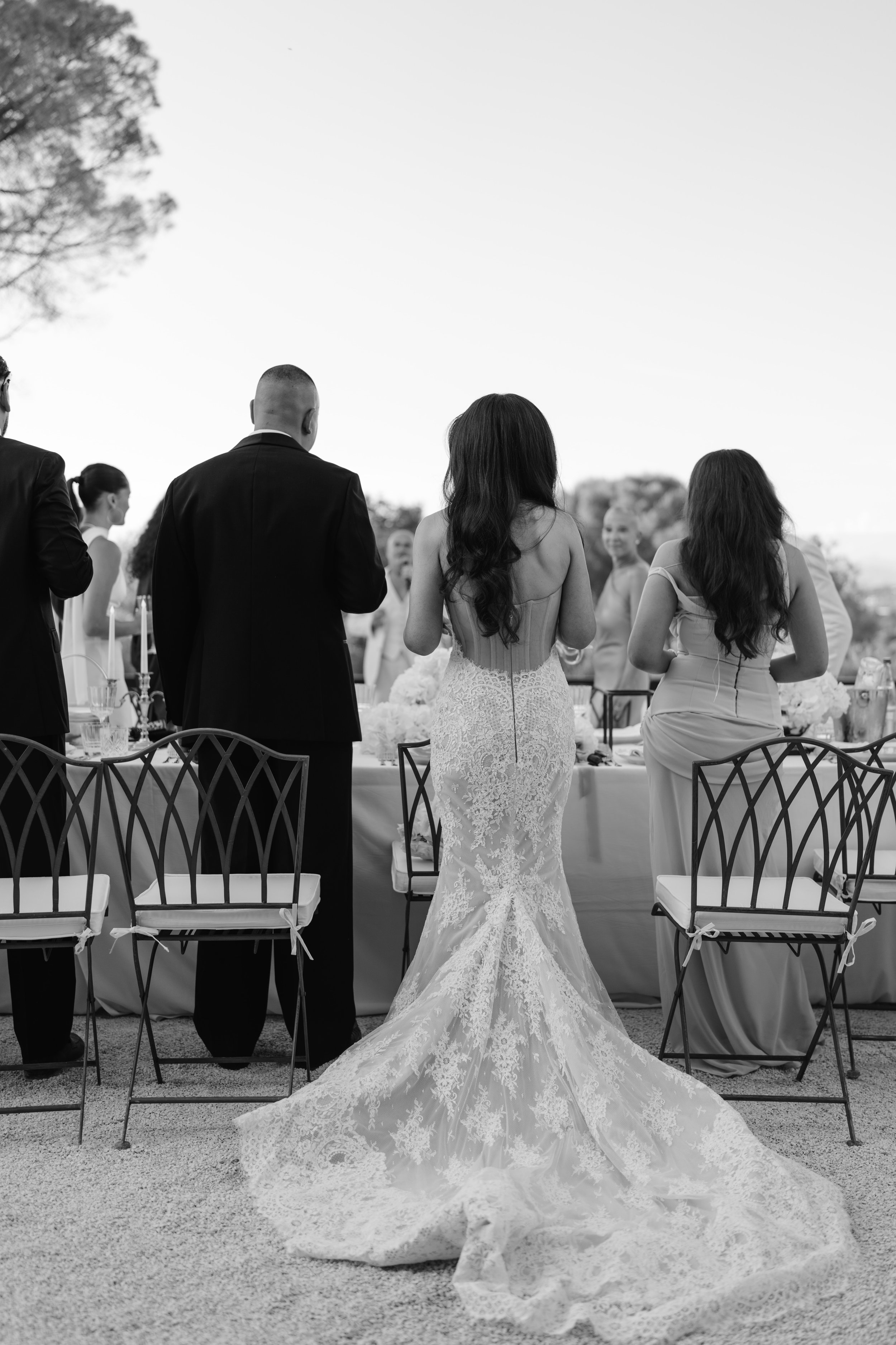 A bride in a lace wedding gown stands with her back to the camera at an outdoor wedding reception, surrounded by seated guests and a decorated table, with the sky and trees in the background.