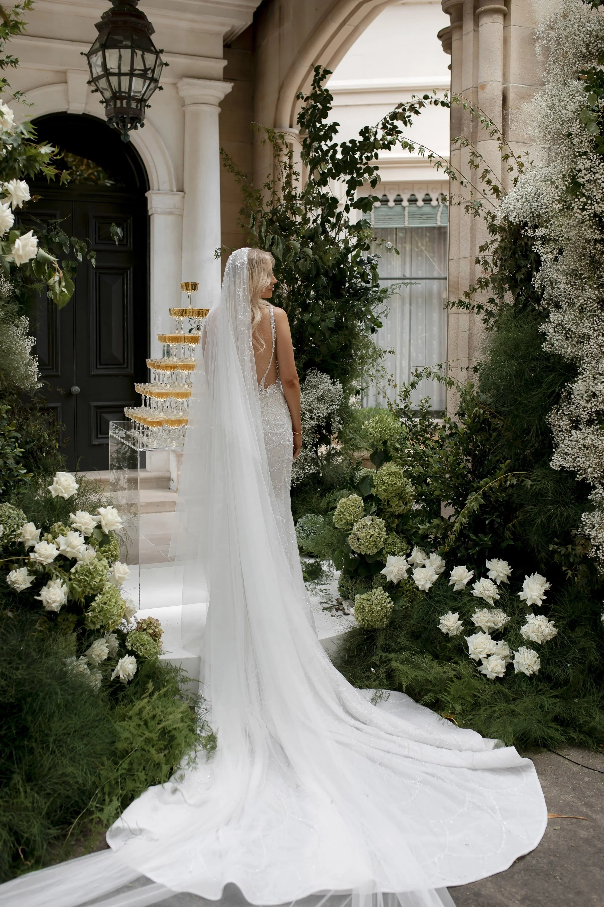 A bride with a long veil surrounded by greenery and a champagne tower.