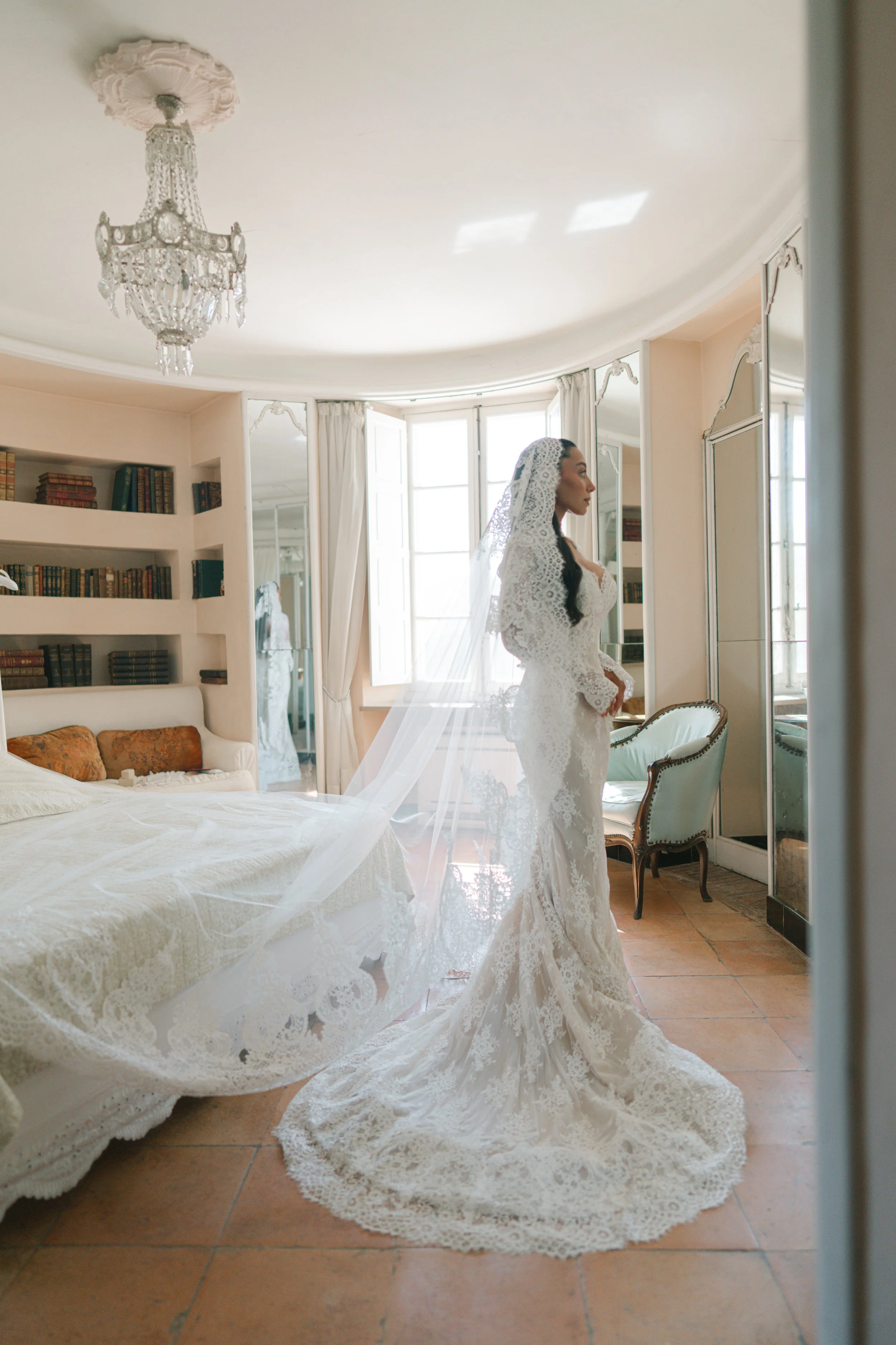 A woman in a wedding dress with a lace veil standing in a room with large mirrors, a chandelier, a bed, and a bookshelf.