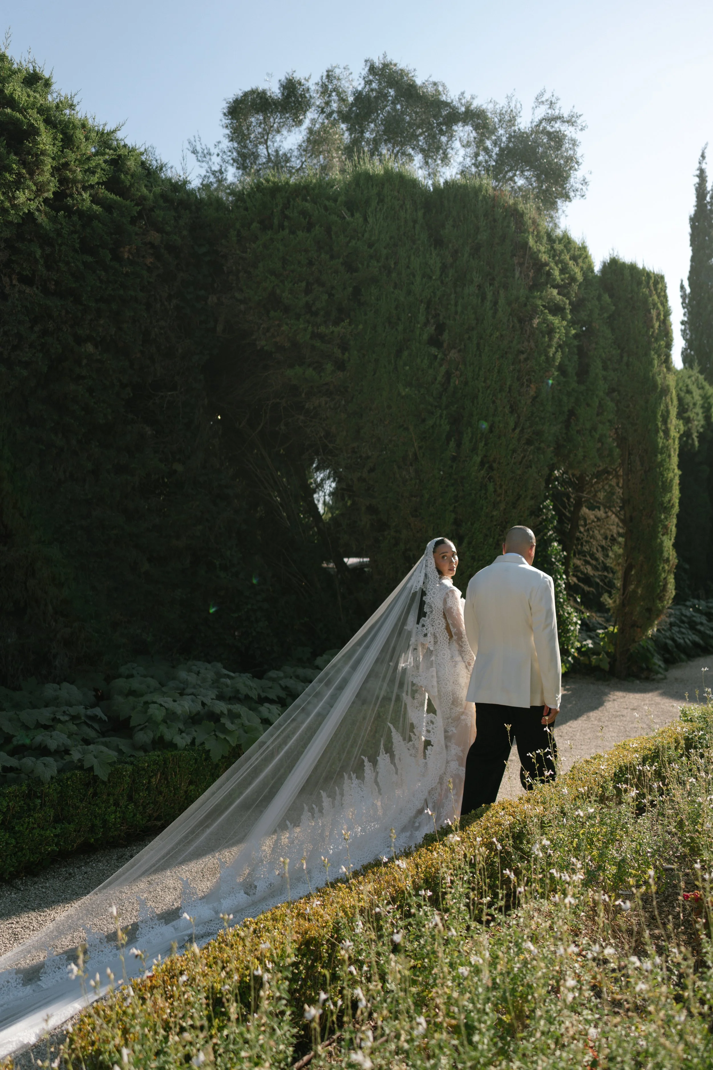 A bride and groom walk through a garden with lush greenery, tall trees, and sunny sky.