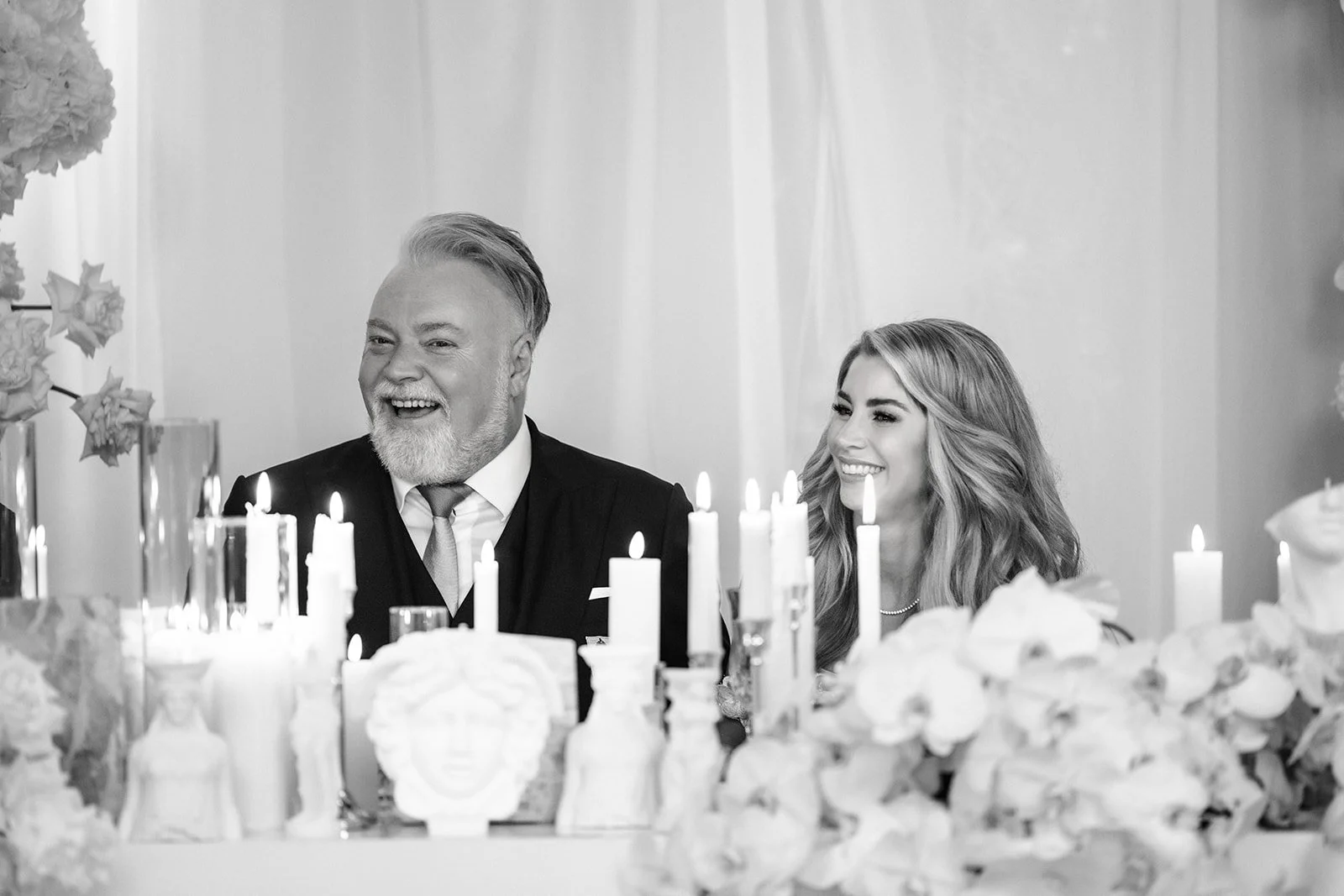 Black and white photo of two people sitting at a table with lit candles and floral decorations, smiling and enjoying a celebration.