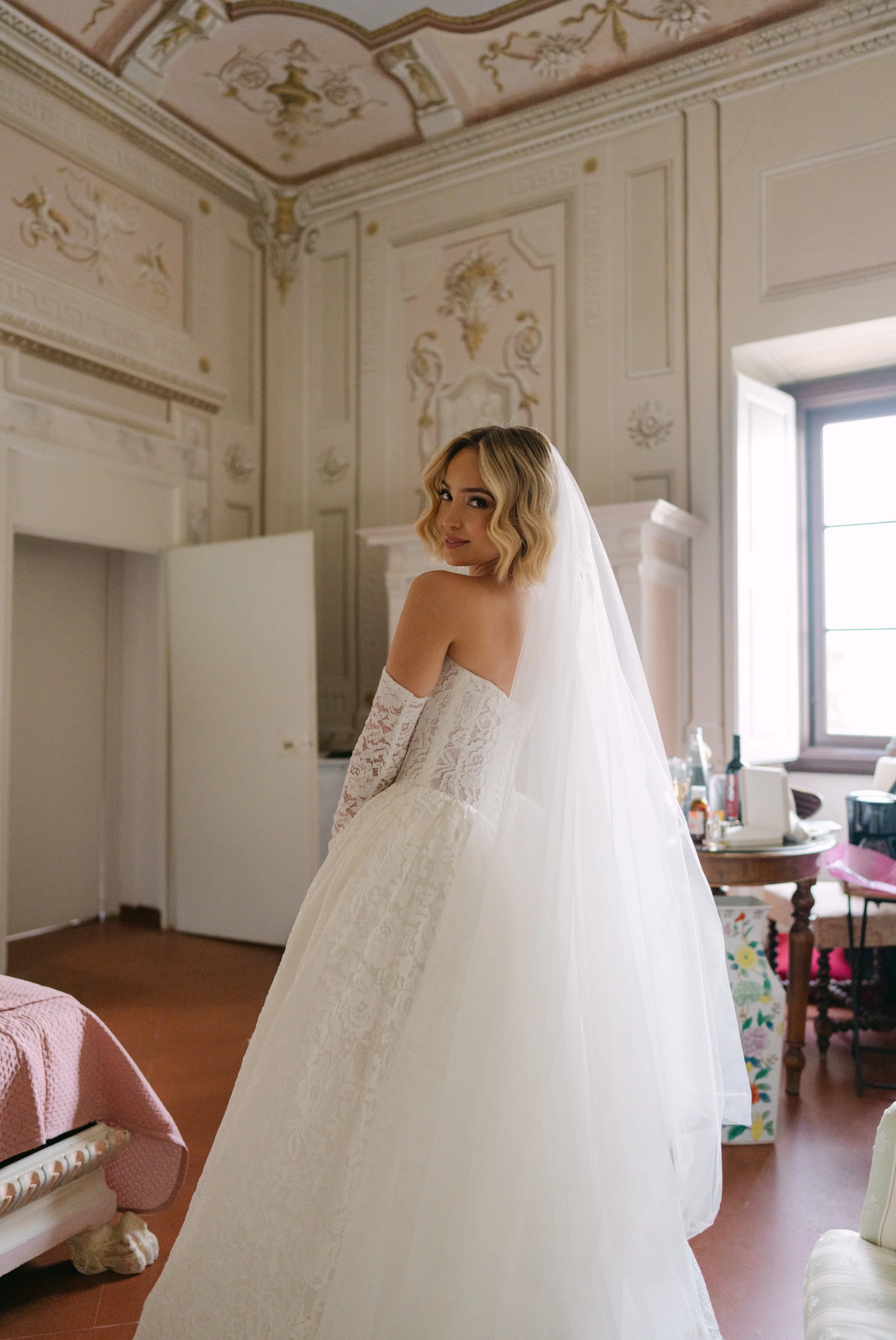 A woman in a wedding dress standing in a decorated room with ornate walls and a window, smiling at the camera.