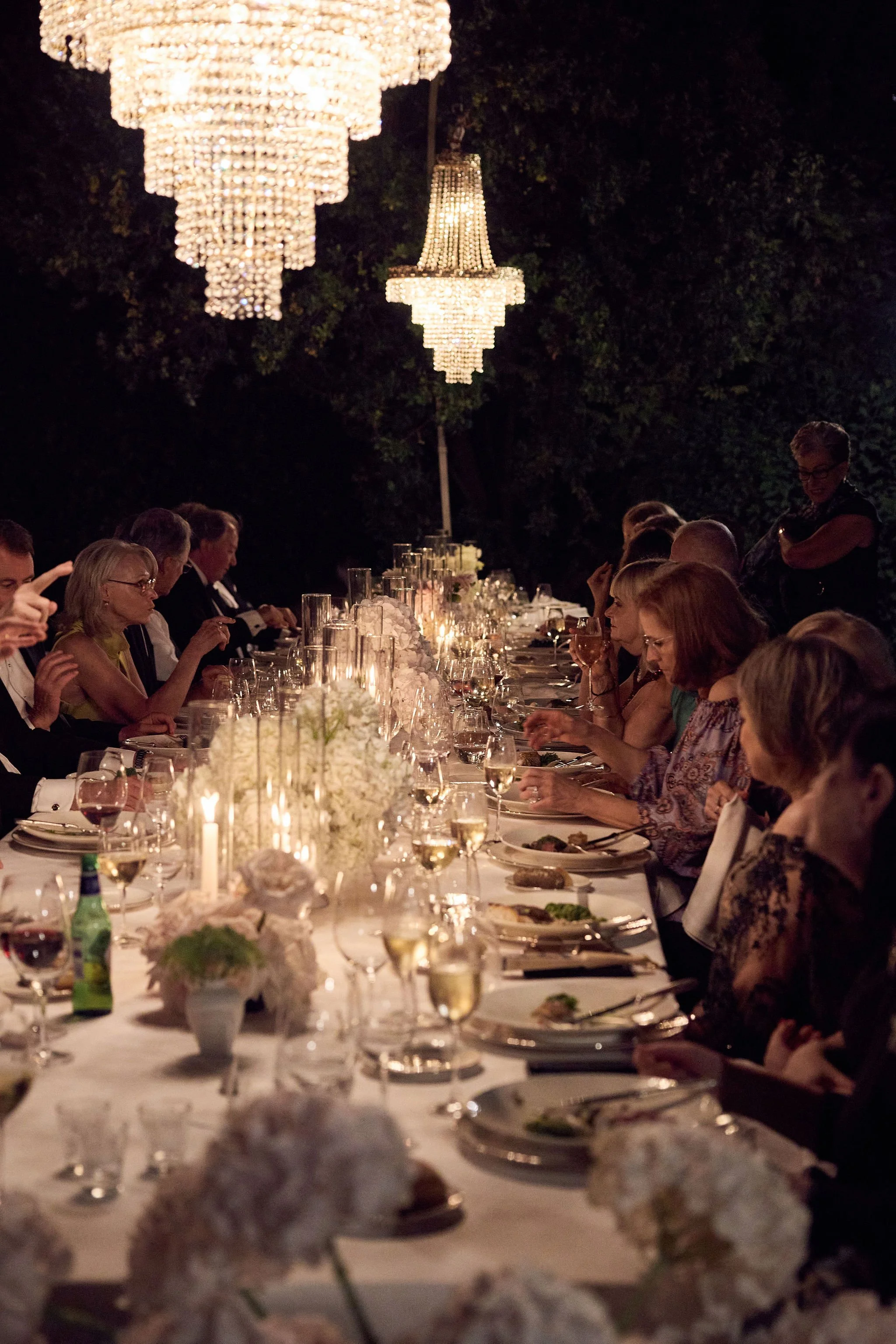 A formal dinner party outdoors at night with a long table, elegant chandeliers overhead, and guests dining with wine glasses and floral centerpieces.