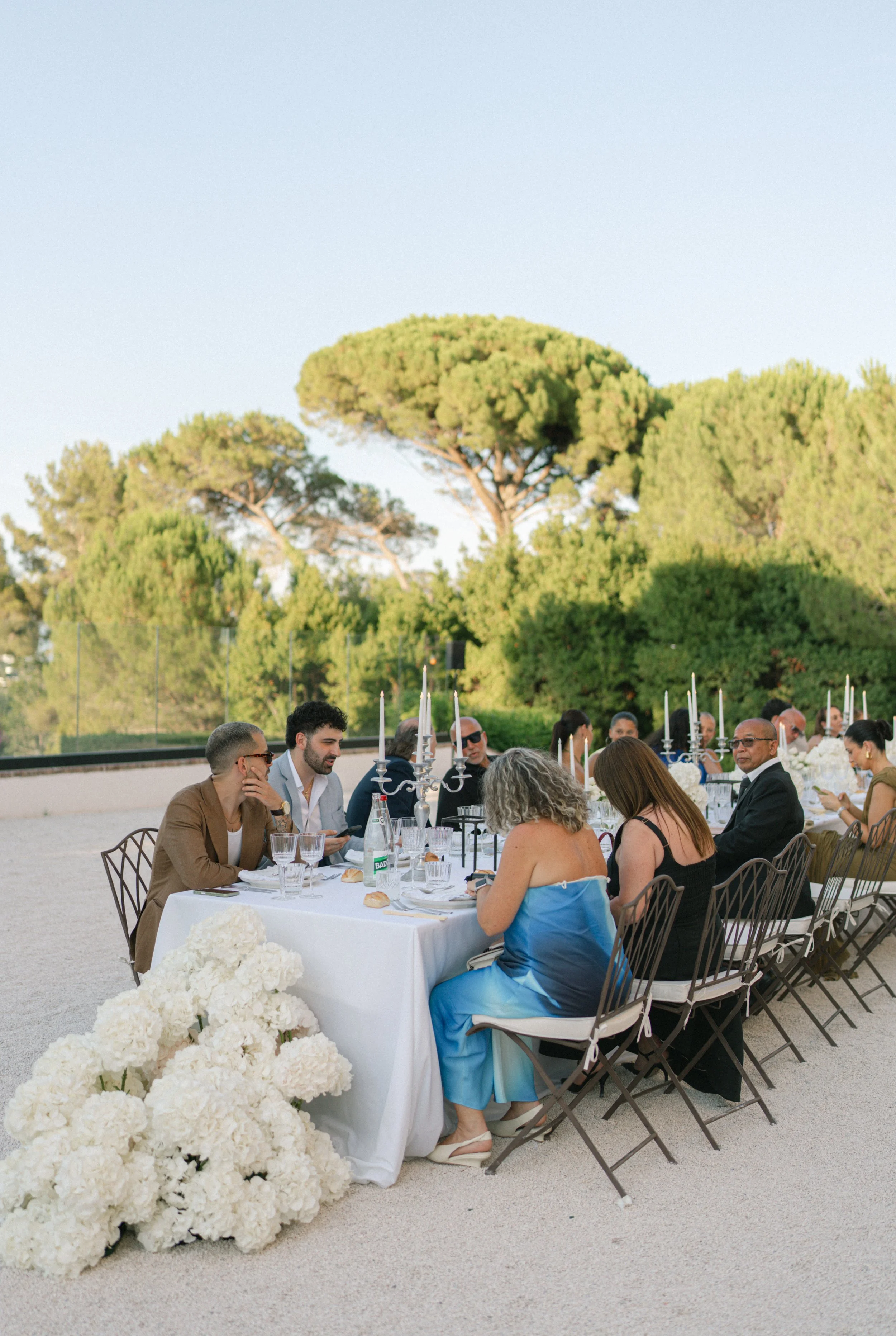 People dining at an outdoor banquet table with white tablecloths and floral centerpieces, surrounded by green trees and a clear sky.
