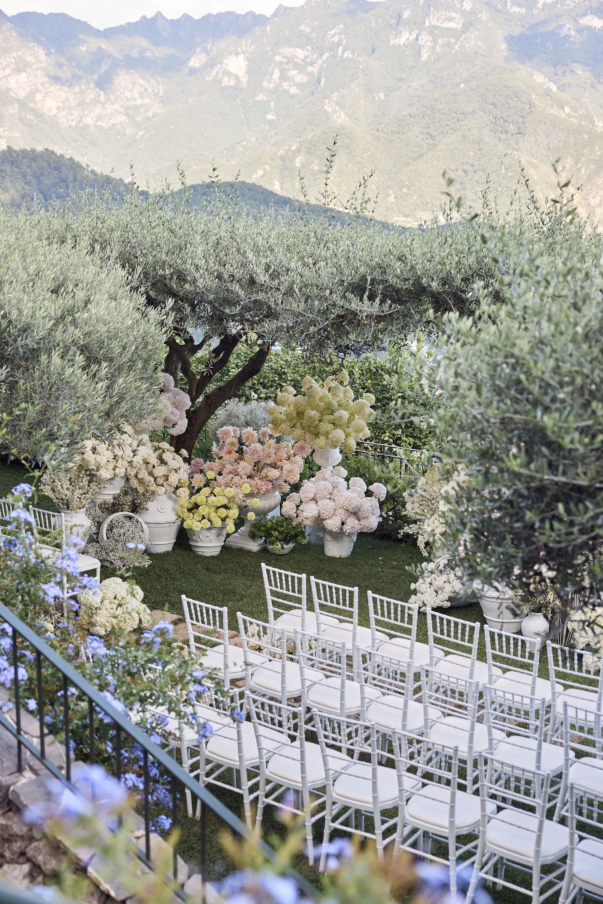 Outdoor wedding setup with white chairs arranged on green grass, surrounded by large flower arrangements in white pots with pink and white flowers, set against lush green trees and mountainous landscape in the background.