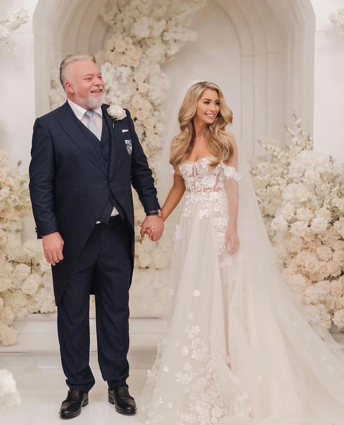 A bride and an older man, likely her father, holding hands during a wedding ceremony in a decorated indoor venue with white flowers.