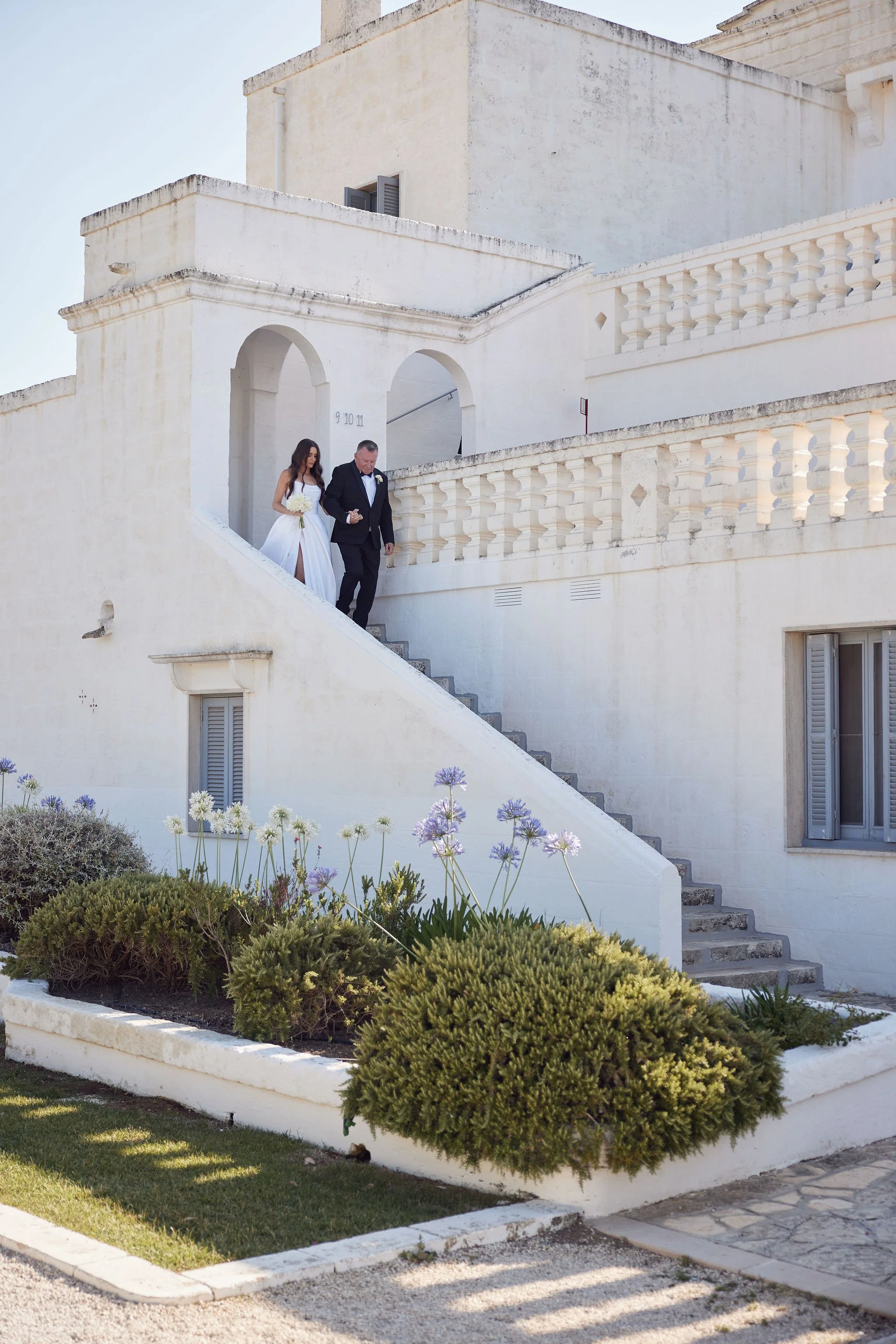 Bride and groom descending a white staircase outside a stucco building, with flowers in the foreground.