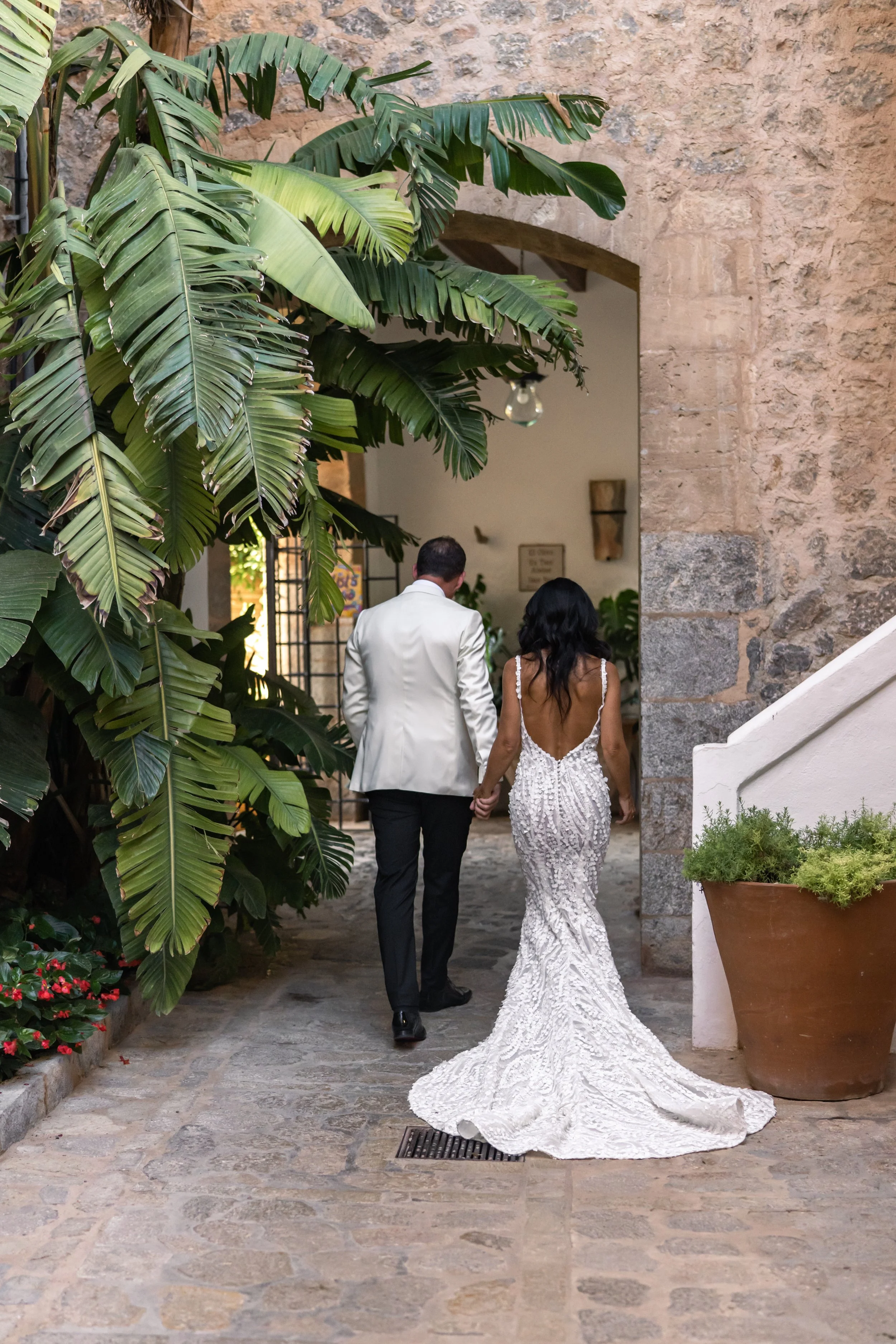 A newly married couple, with the woman in a white wedding gown and the man in a white tuxedo jacket, holding hands as they walk through an archway in a rustic courtyard filled with lush green plants.