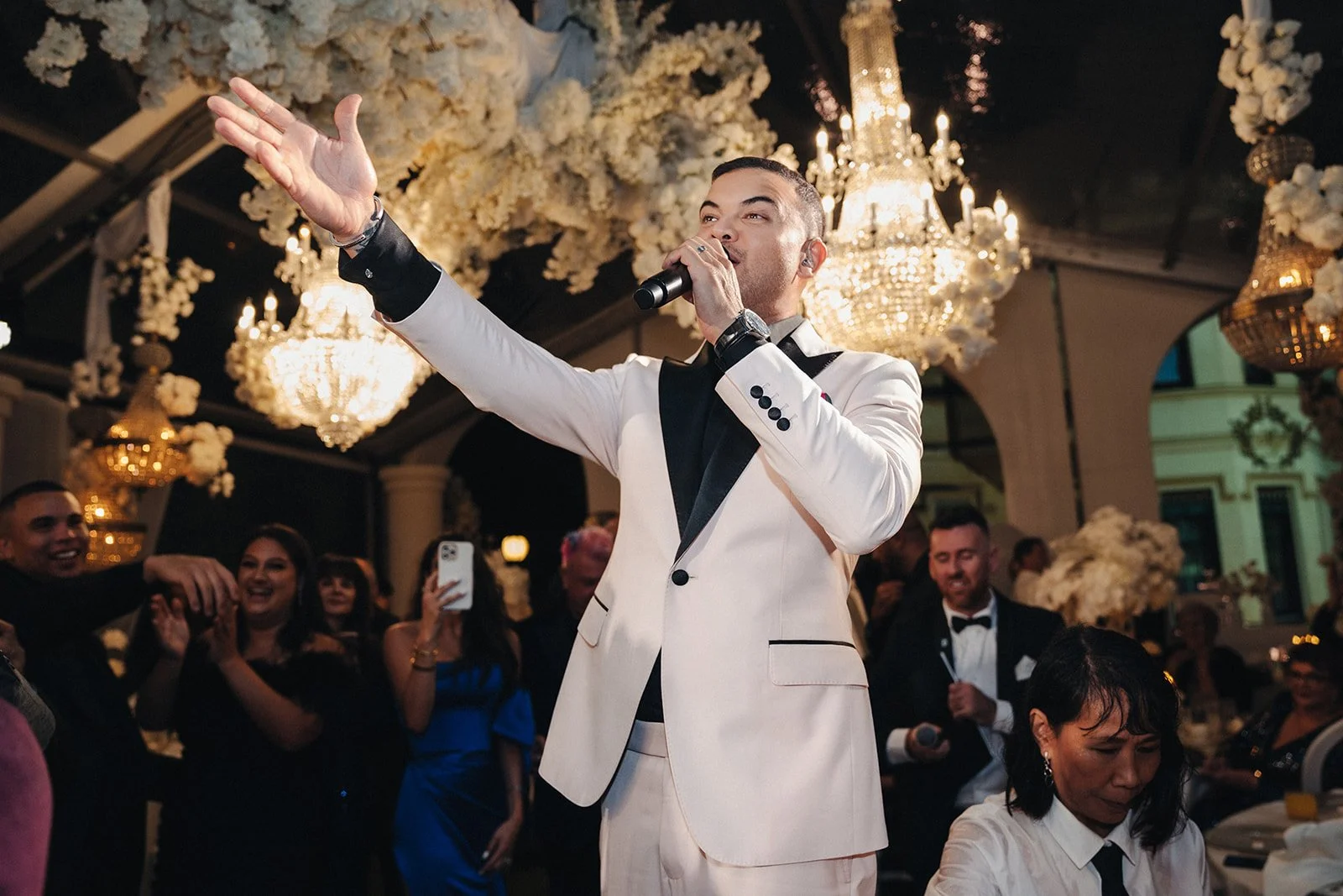 A man in a white tuxedo with black lapels singing into a microphone at a formal event, surrounded by a crowd of people in elegant attire, with ornate chandeliers and floral decorations overhead.