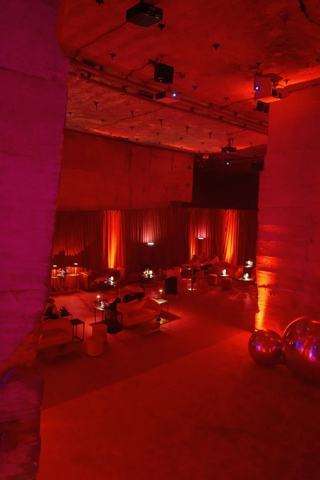 Dimly lit lounge with red lighting, couches, small tables with flowers and lamps, and people sitting and talking.