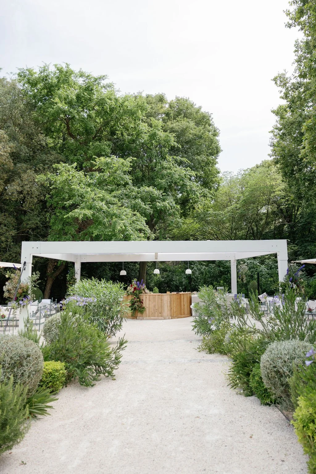 Outdoor event space with a white pergola, surrounded by green trees and plants, with a wooden bar and floral arrangements.