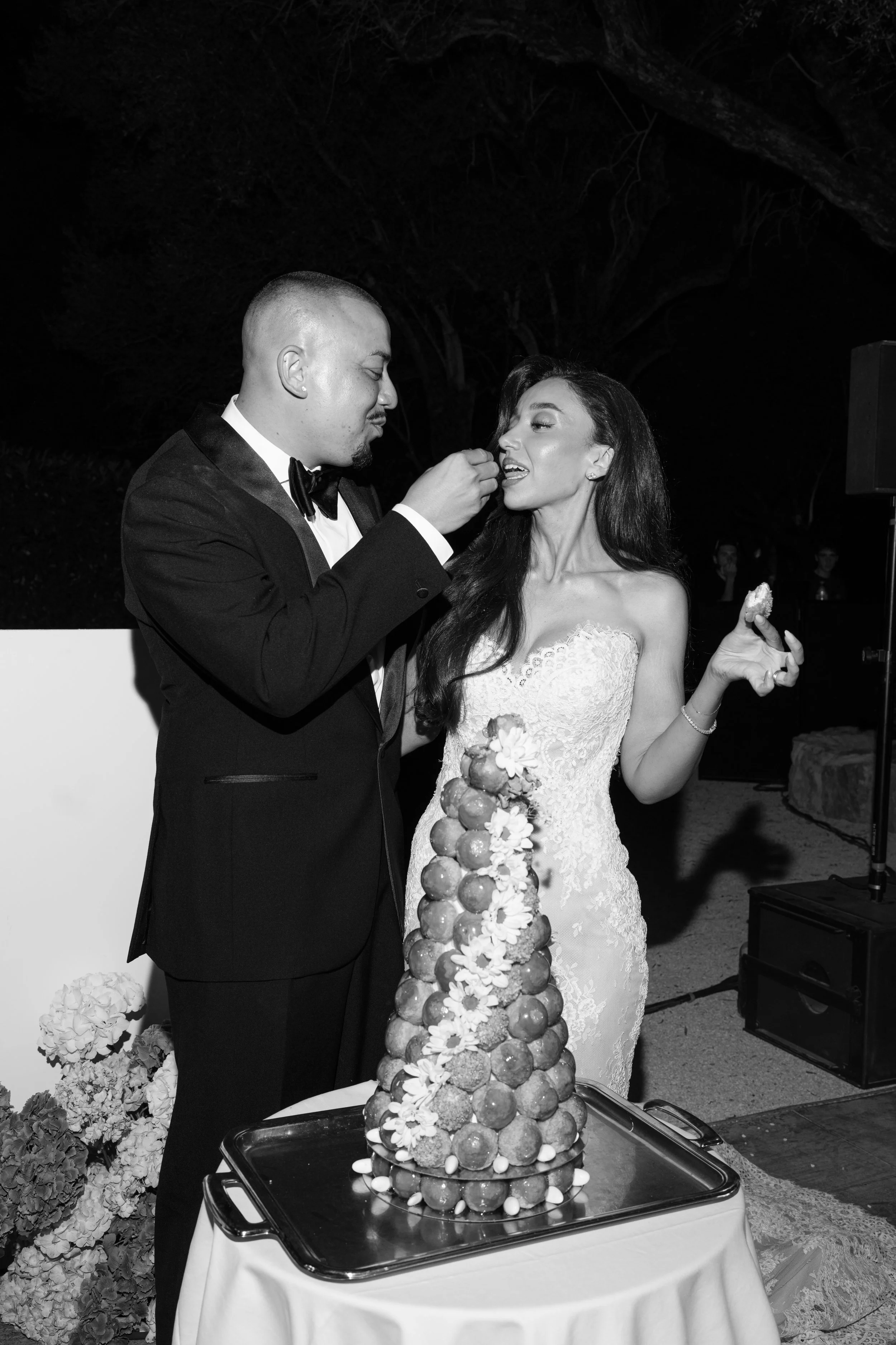 A black and white photo of a wedding celebration with a bride and groom. The groom in a tuxedo is feeding the bride, who is wearing a wedding dress, from a tall, layered wedding cake decorated with fruits and flowers. They are outdoors at night, with