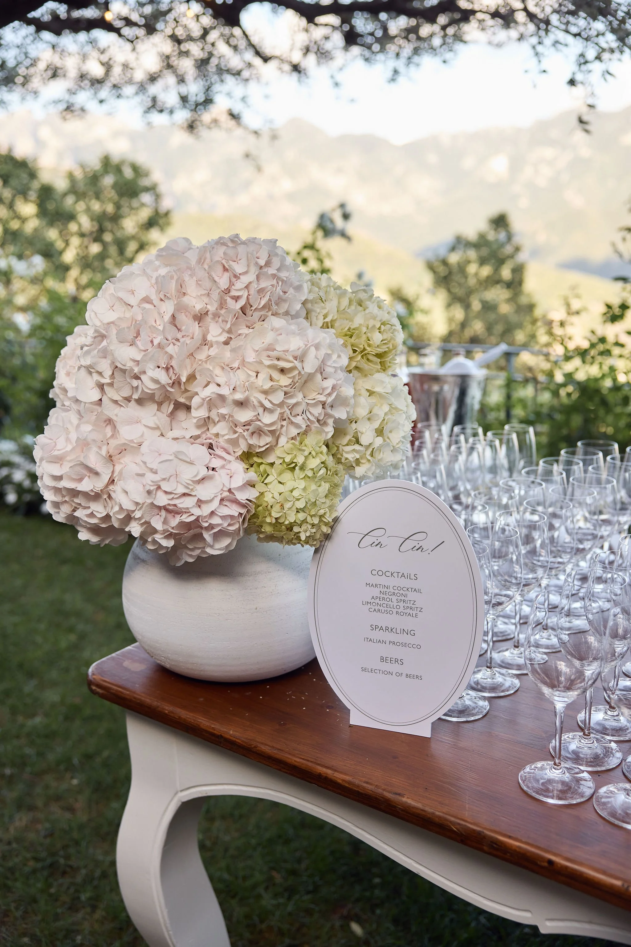 A large bouquet of pale pink and white hydrangeas in a white vase on a wooden table, with a sign listing drinks and alcohol options, and a background of a lush, green outdoor setting.