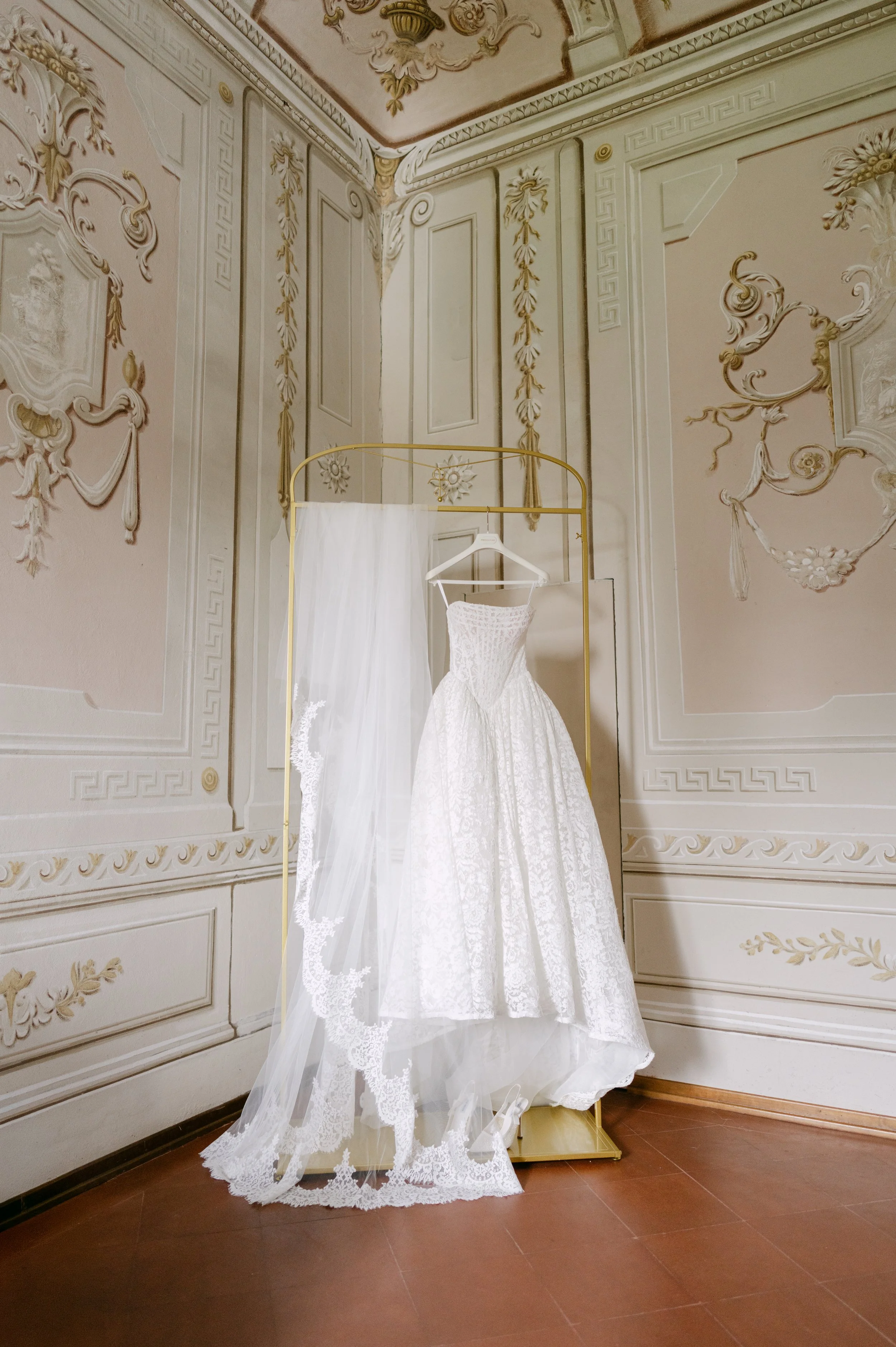 A white wedding dress with lace details hanging on a gold clothing rack in a room with ornate, rococo-style decorated walls and ceiling in soft pink, beige, and gold tones.