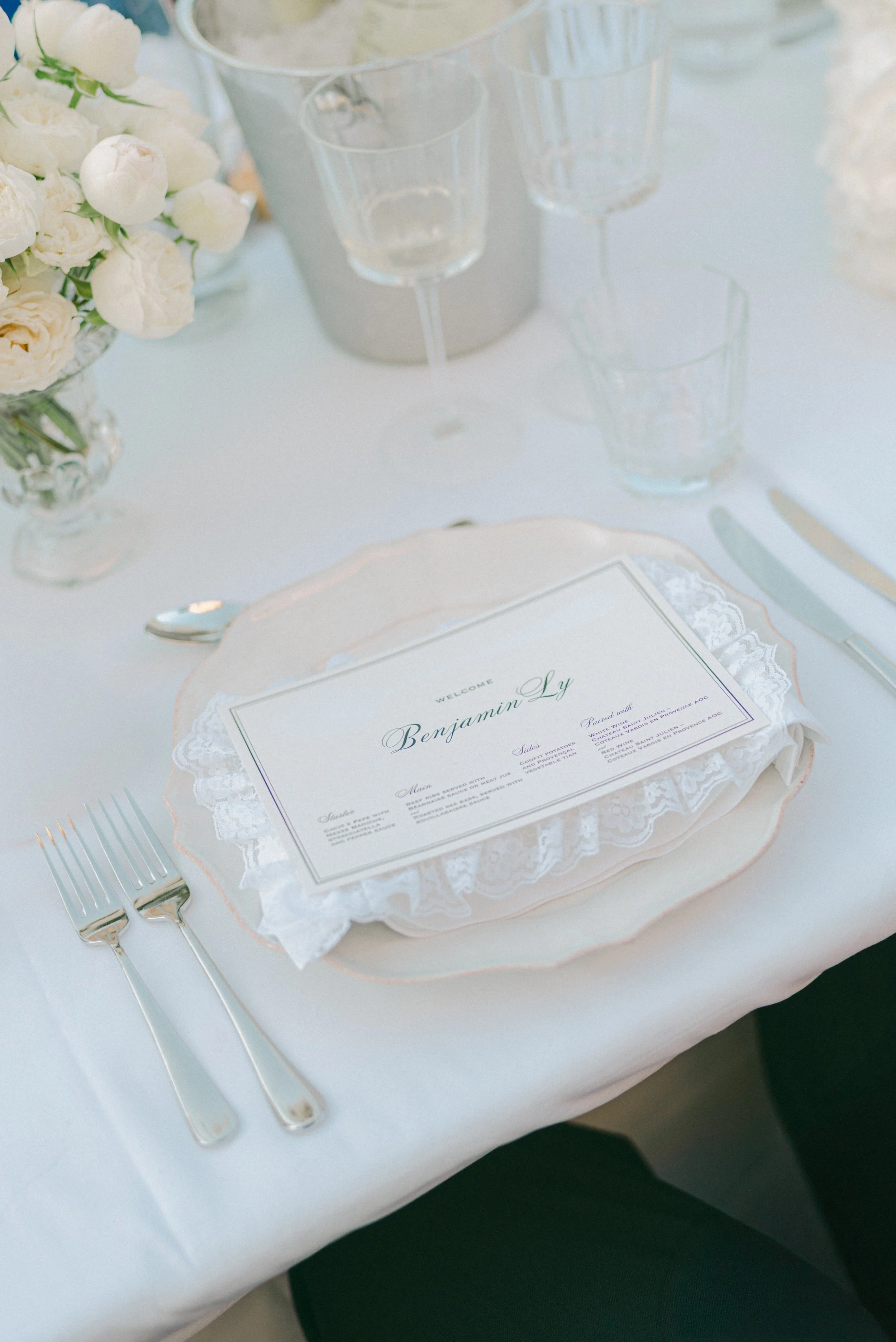 A wedding reception table setting with a menu card, two forks, and a vase of white flowers.