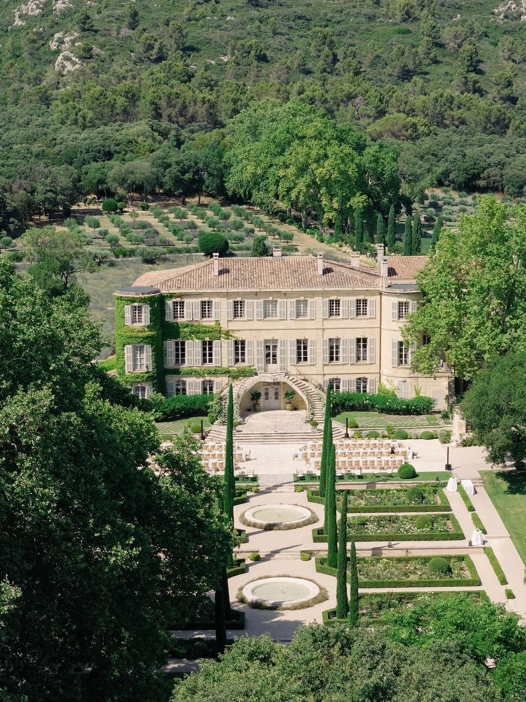 A large, elegant house surrounded by manicured gardens and tall trees, set against a hillside with terraced plantings.