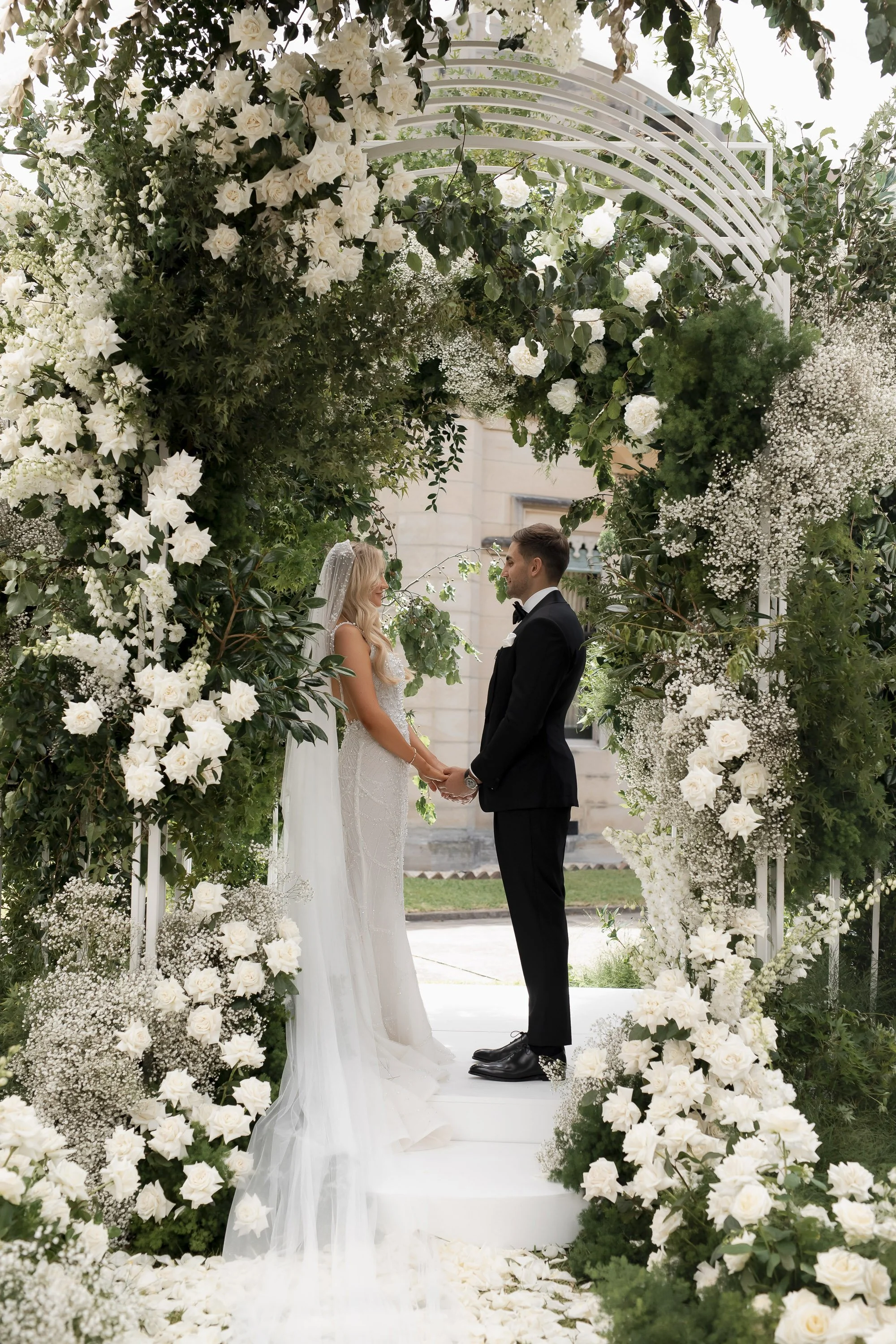 A bride and groom holding hands and facing each other during a wedding ceremony outdoors, framed by a floral arch filled with white flowers and greenery.