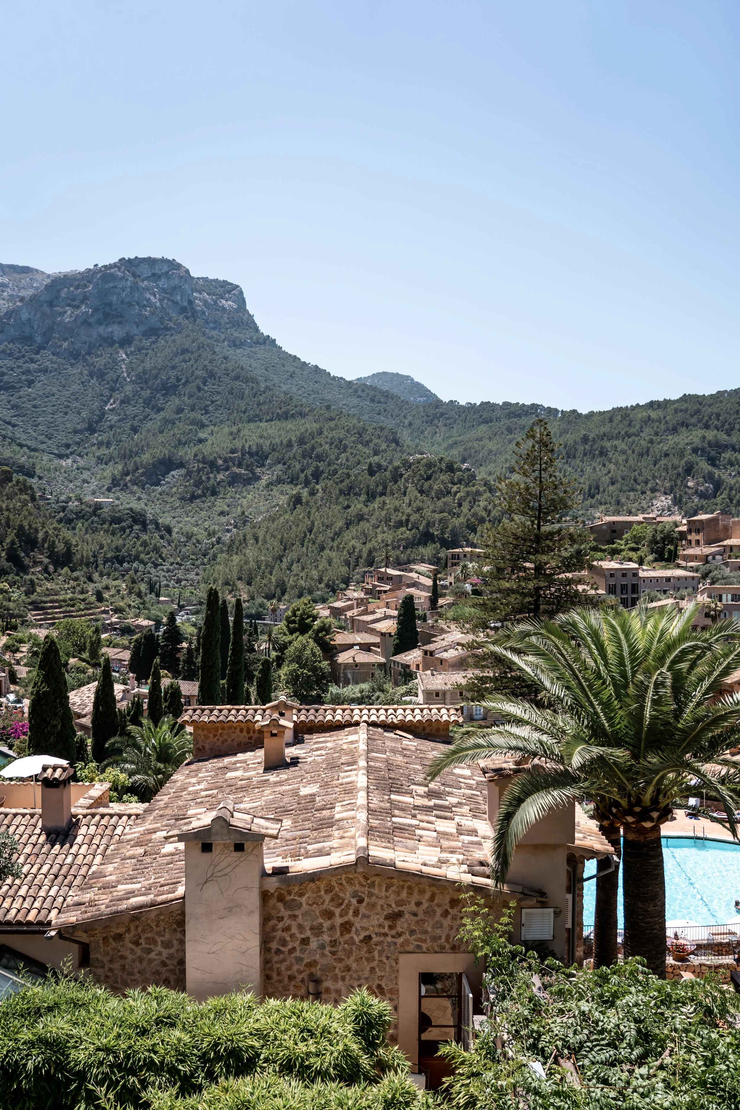 Tiled rooftops and trees overlooking a hillside town and surrounding mountains.