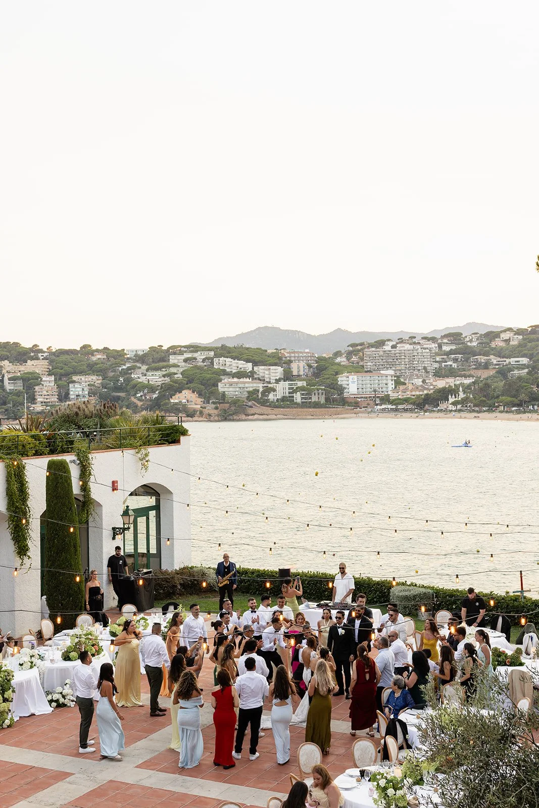 Outdoor wedding reception with guests dancing and live band by a waterfront with hilly residential area in the background.
