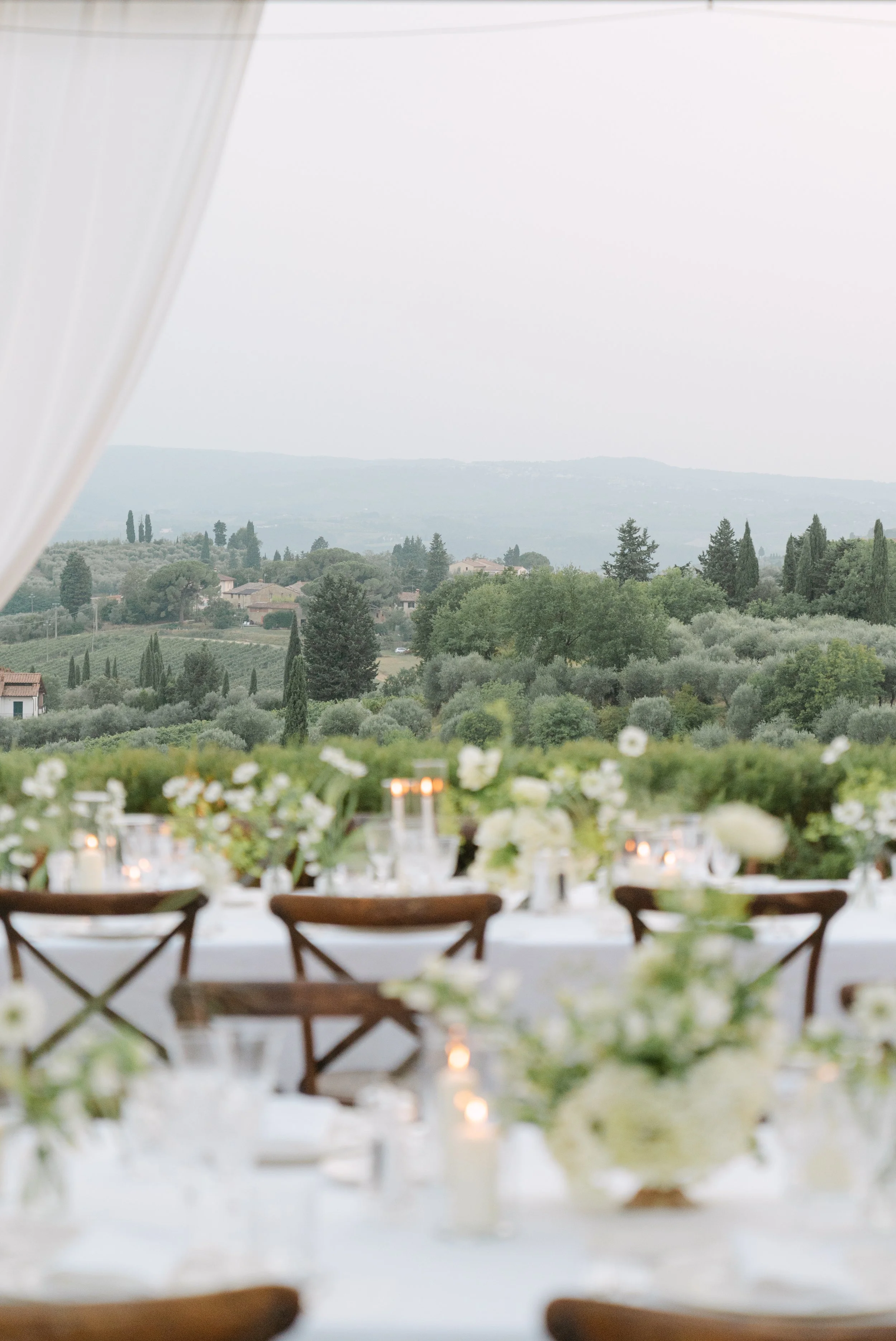 Indoor dining table set with candles and floral arrangements, overlooking a scenic landscape of trees and distant hills through large windows.
