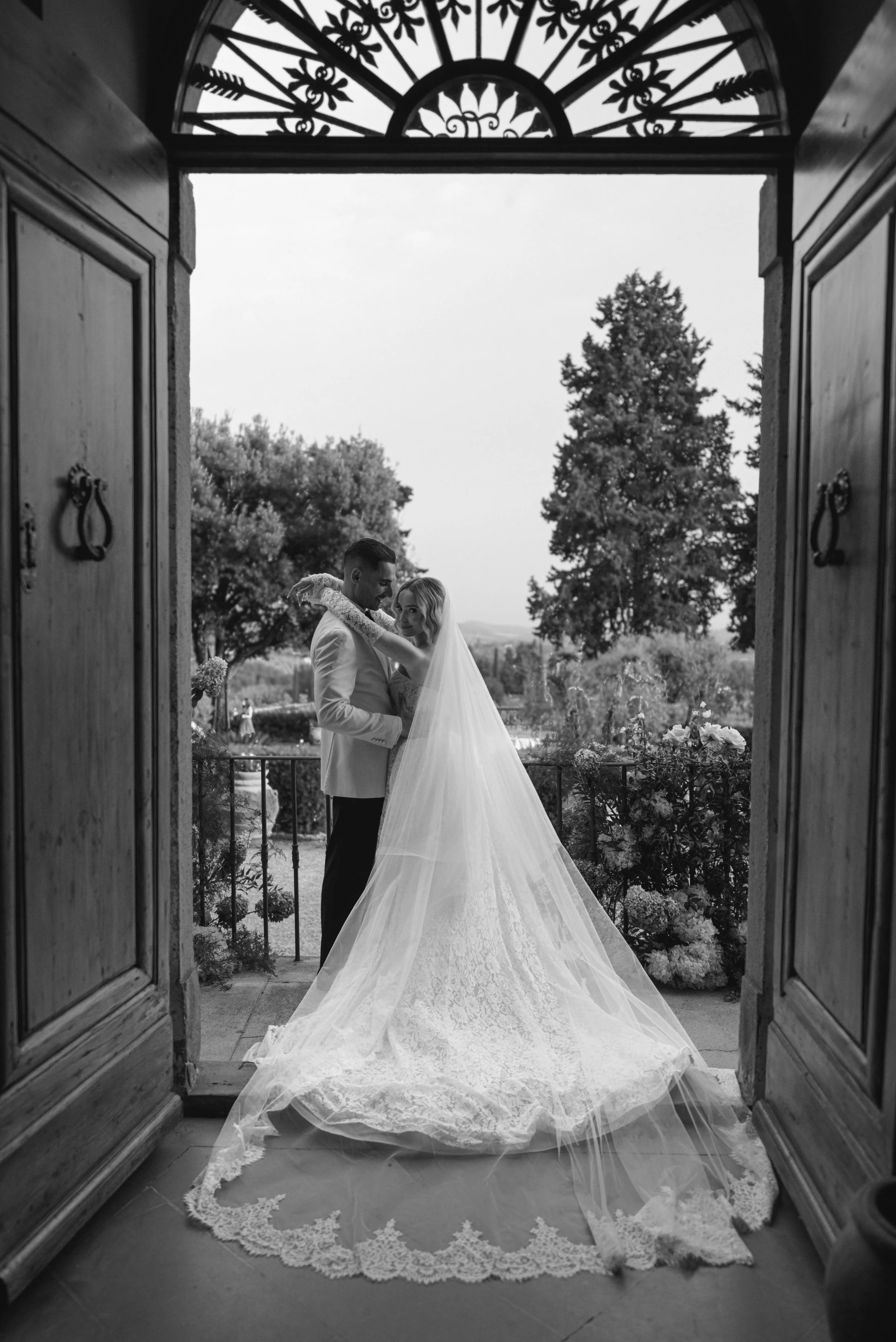 A couple in wedding attire stands in an open doorway, with the bride in a long lace dress and veil, and the groom in a white suit. The scene captures a moment of intimacy, with trees and a landscape visible in the background. The photo is black and w
