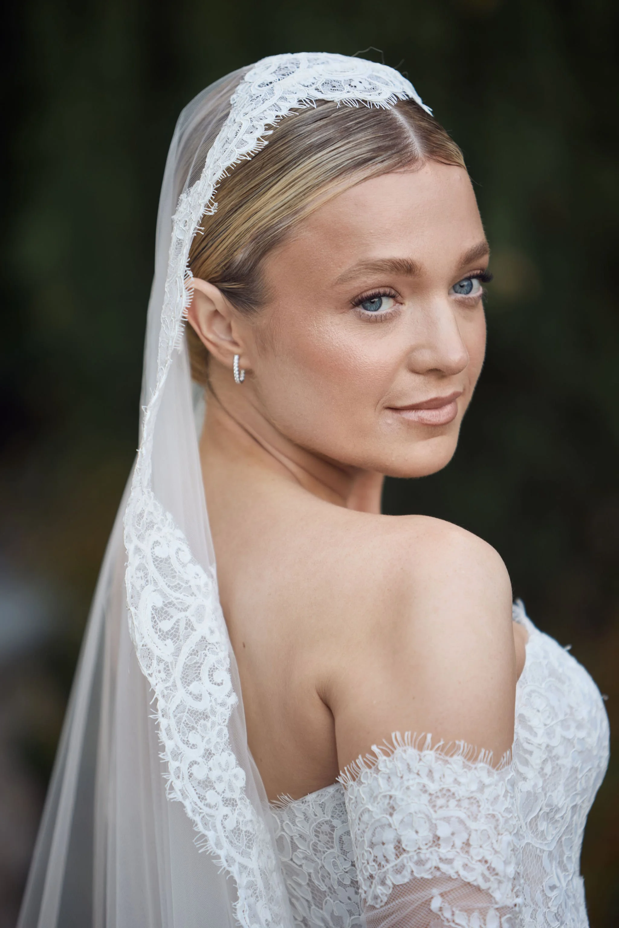 Close-up of a bride with blonde hair, blue eyes, wearing lace wedding dress, veil, and earrings, looking over her shoulder with a soft smile.