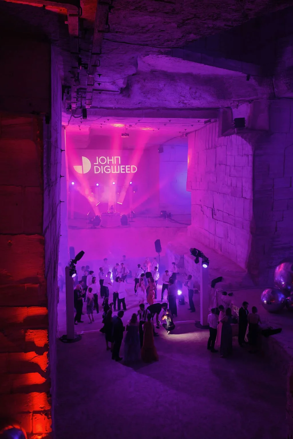 An indoor party or concert with purple lighting and people dancing and socializing, with DJ John Digweed's name projected on the wall in the background.