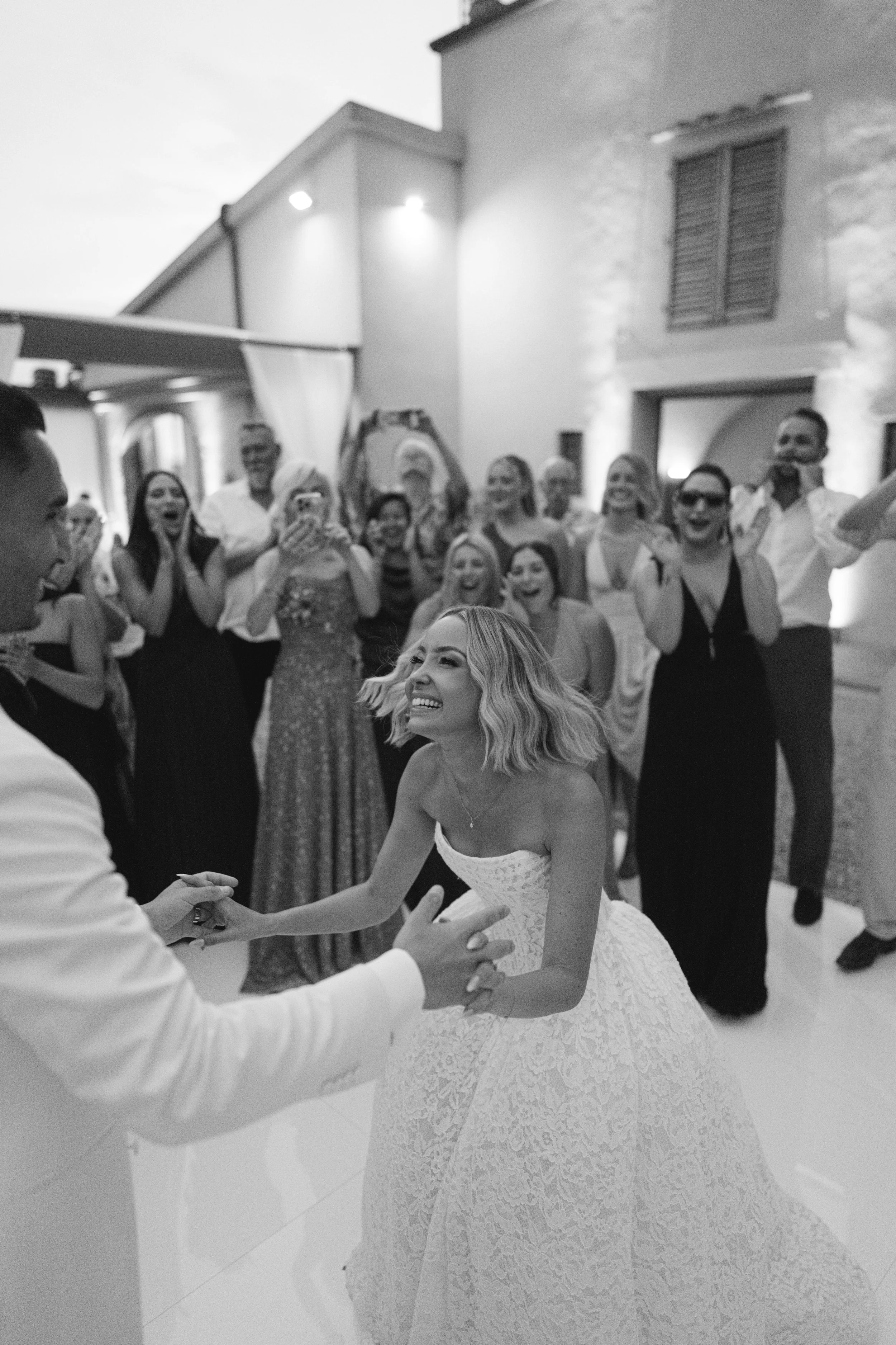 A bride and groom are dancing at their wedding reception, with guests in the background watching and smiling.